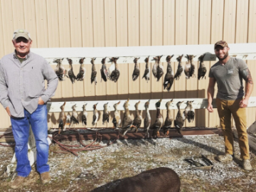 Two men posing with a long row of hanging hunted birds in front of a beige building
