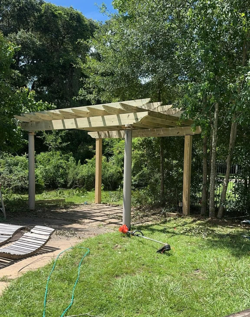 Wooden pergola in a grassy garden surrounded by trees and shrubs, with a net lying on the ground.