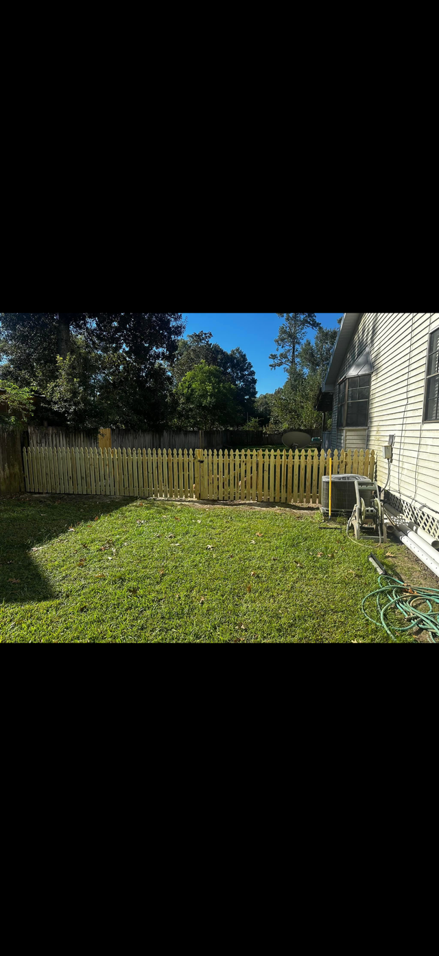 Small fenced backyard with green grass, wooden fence, trees, and a house wall on the right.