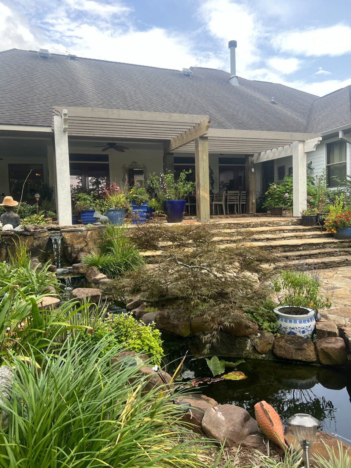 Backyard pond with koi, stone steps, lush plants, and a covered patio under a wavy roof