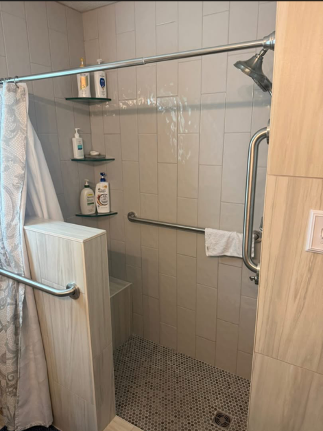 Accessible shower with grab bars, shower chair, handheld showerhead, and built-in shelves with toiletries.