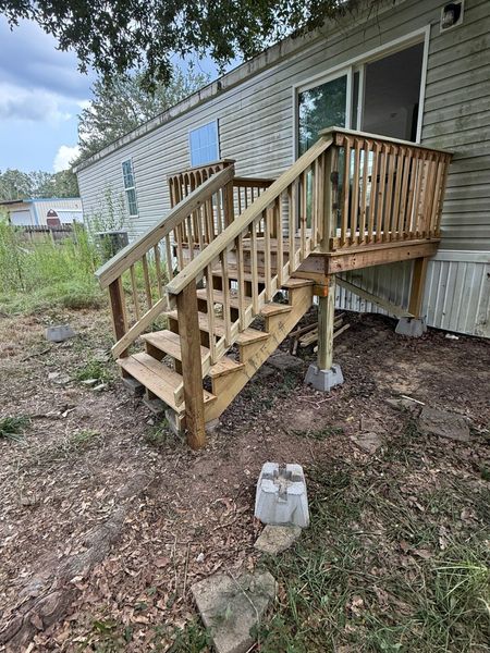 Wooden porch stairs with railings leading to a white house, unfinished and set over gravel and dirt.
