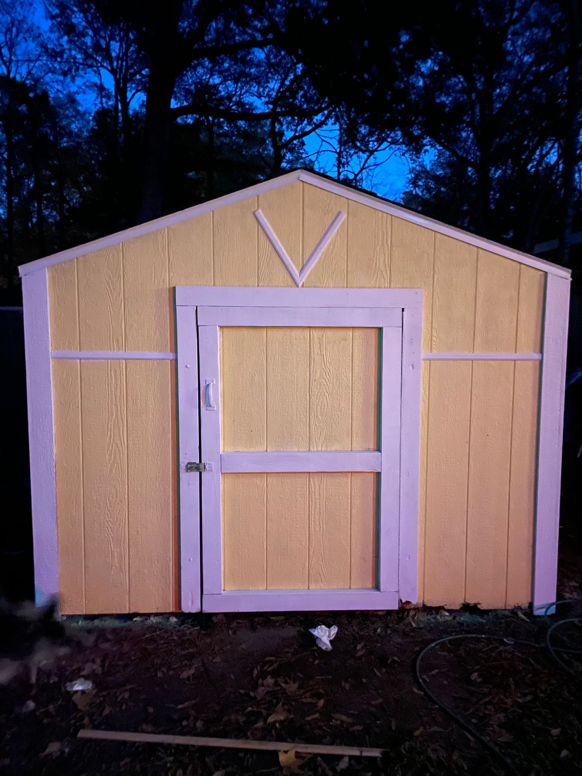 Small tan shed with a pale purple door and trim, set outdoors at dusk