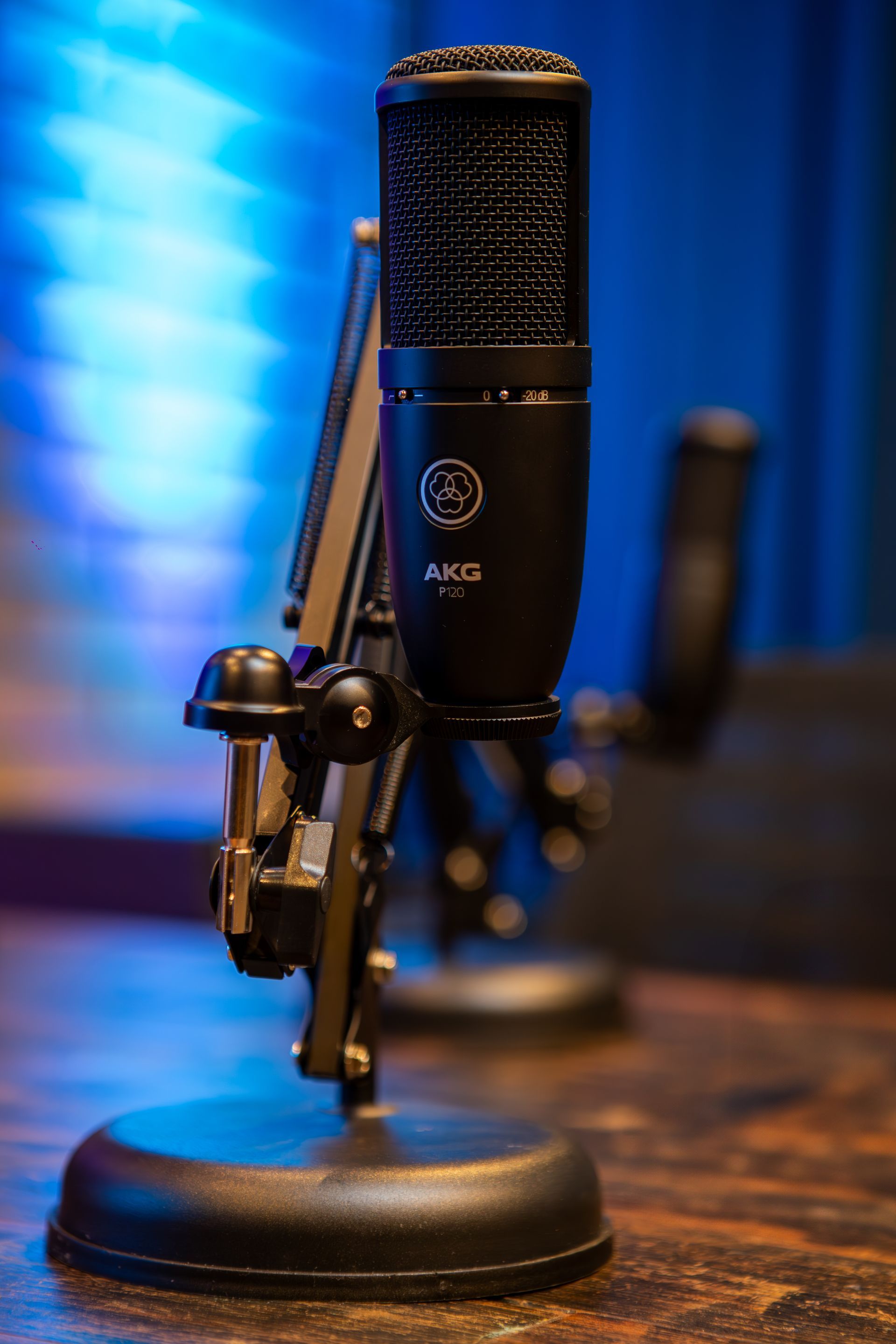 Um microfone AKG preto montado em um suporte, posicionado sobre uma superfície de madeira, com um fundo azul.