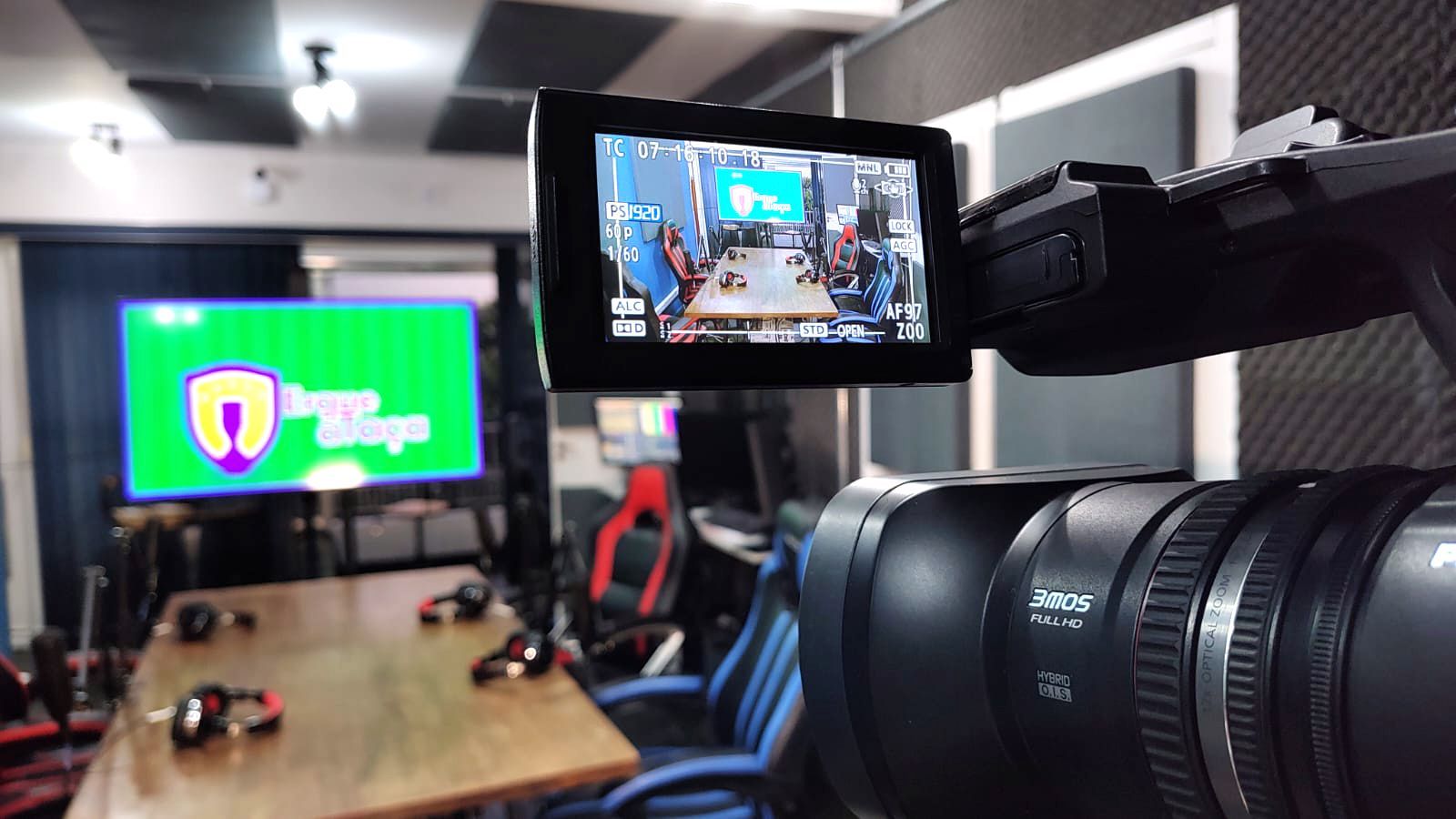 Câmera filmando um estúdio com mesa, cadeiras e uma tela exibindo um logotipo. A tela é verde e roxa.
