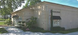 Small beige storefront building with awnings and shrubs on a sunny street corner