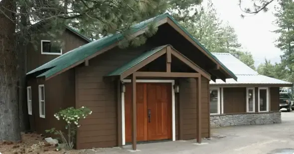 Rustic brown wooden building with green roof and small porch in a wooded area
