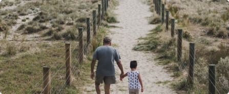 Two people holding hands, walking down a sandy path between fence posts in a grassy field.