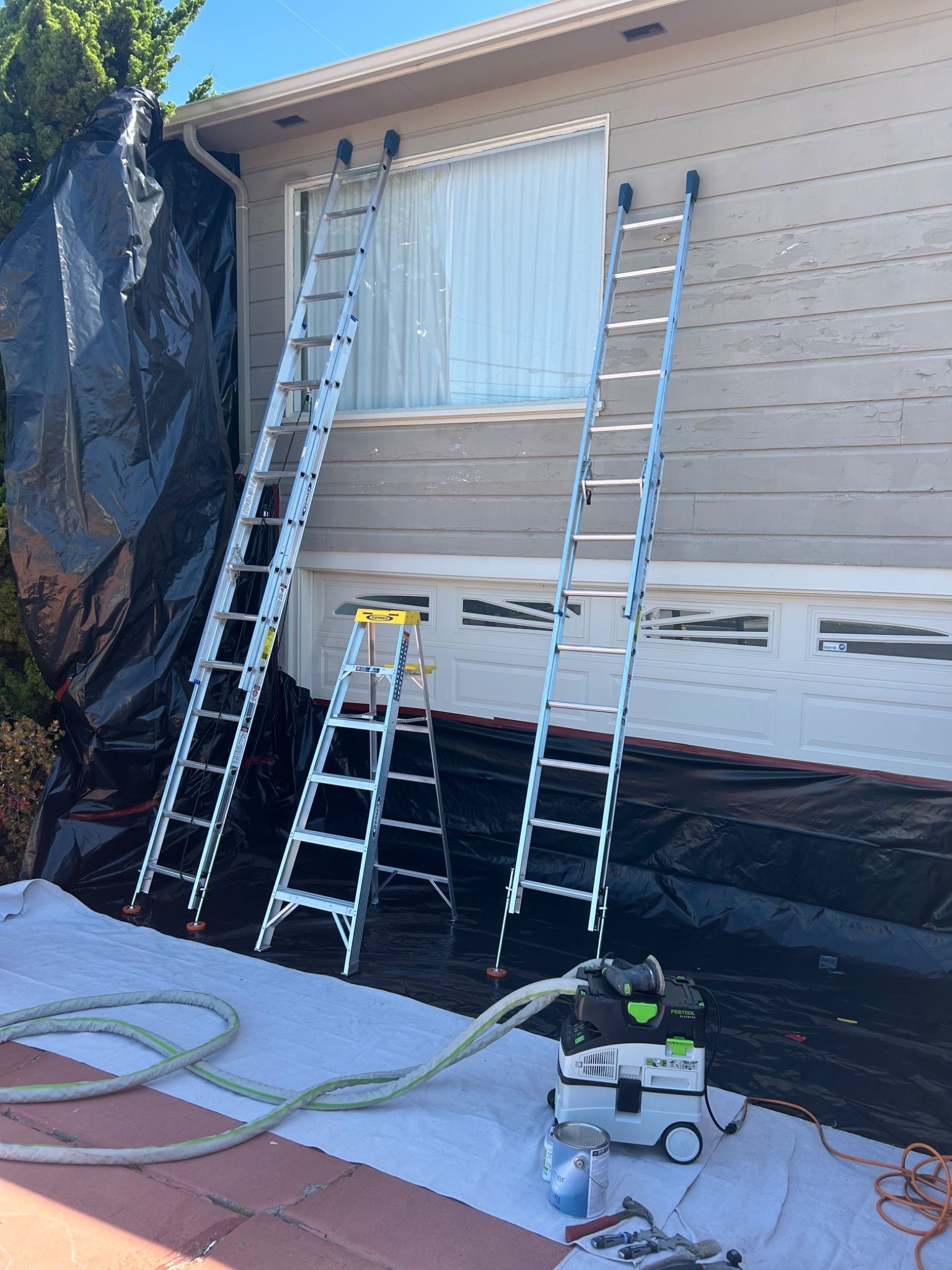 Ladders leaning against a house exterior with a covered window and renovation tools below