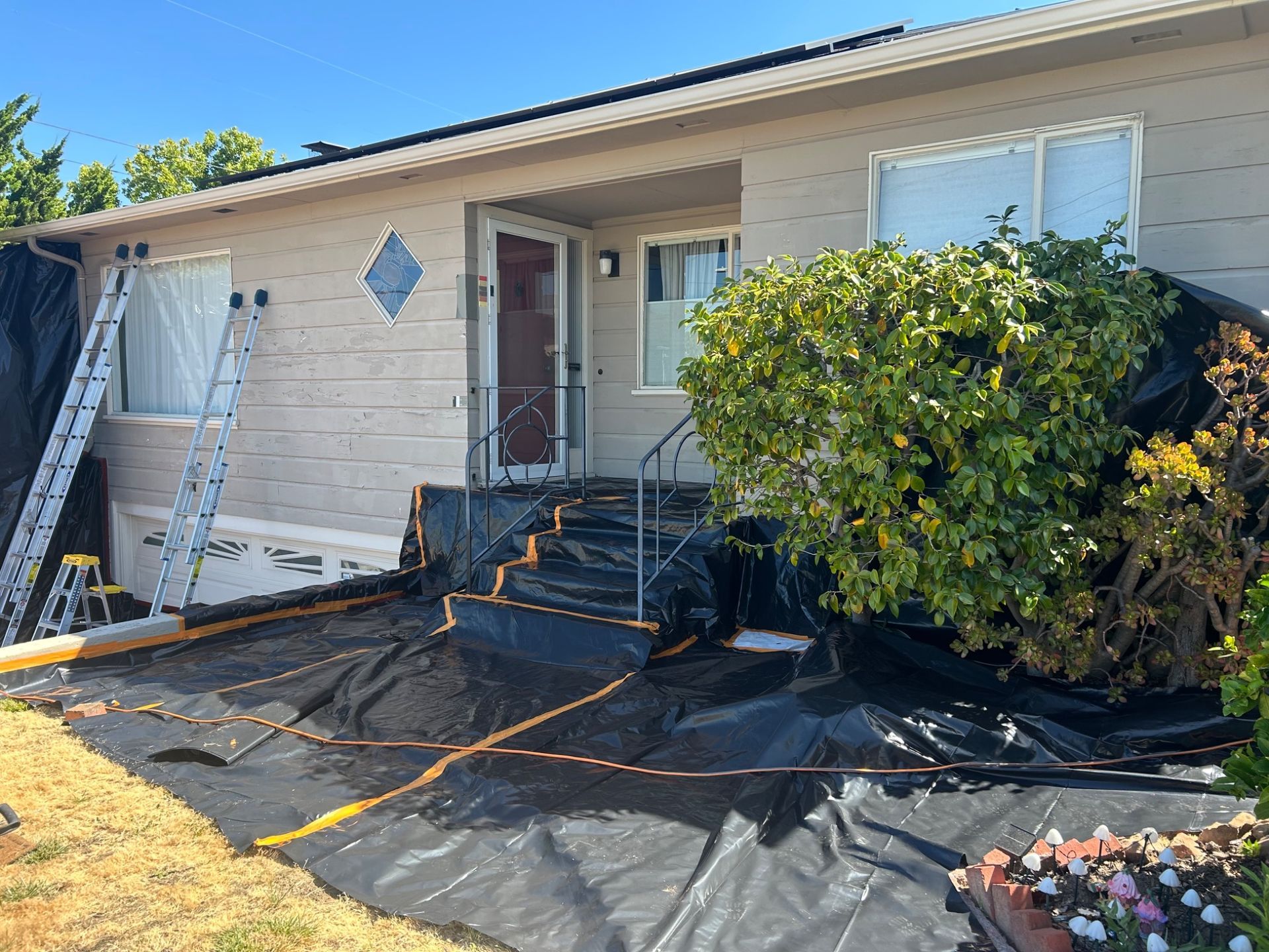Single-story house front porch with ladder, black tarp ground cover, and bushes in front yard