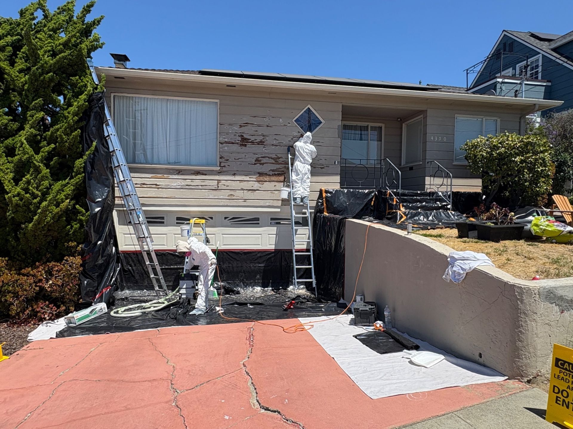 Workers repairing a cracked red driveway outside a house with wood siding and a white gate