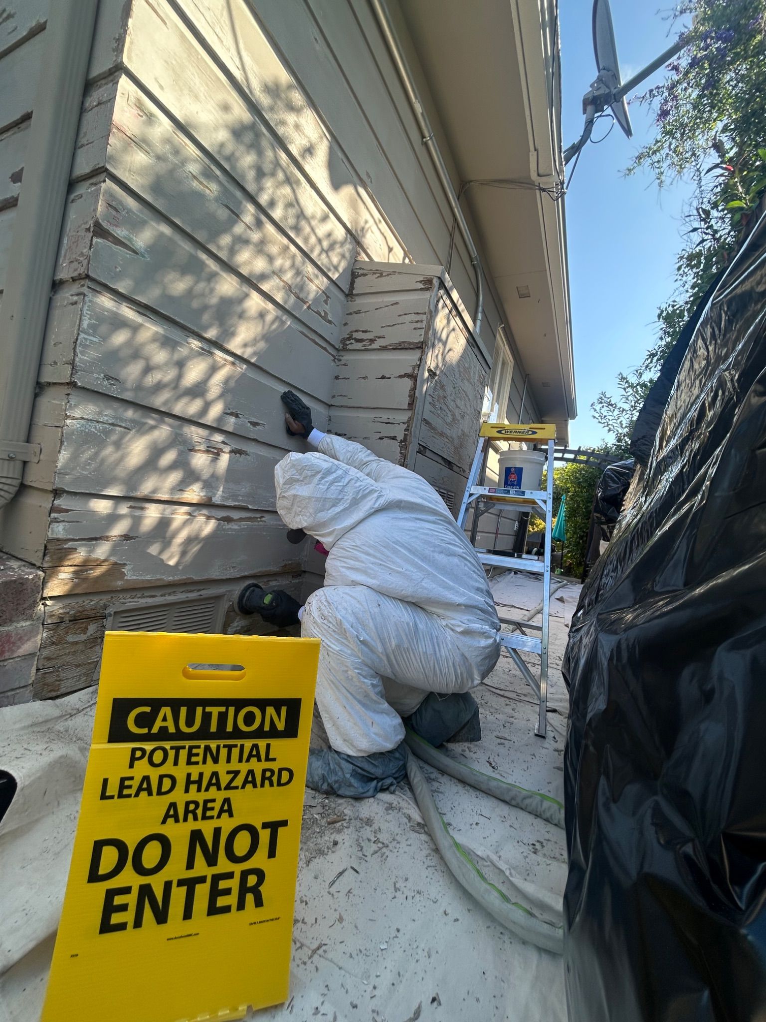 Worker in white protective suit inspecting house wall near a yellow “Do Not Enter” lead hazard sign.