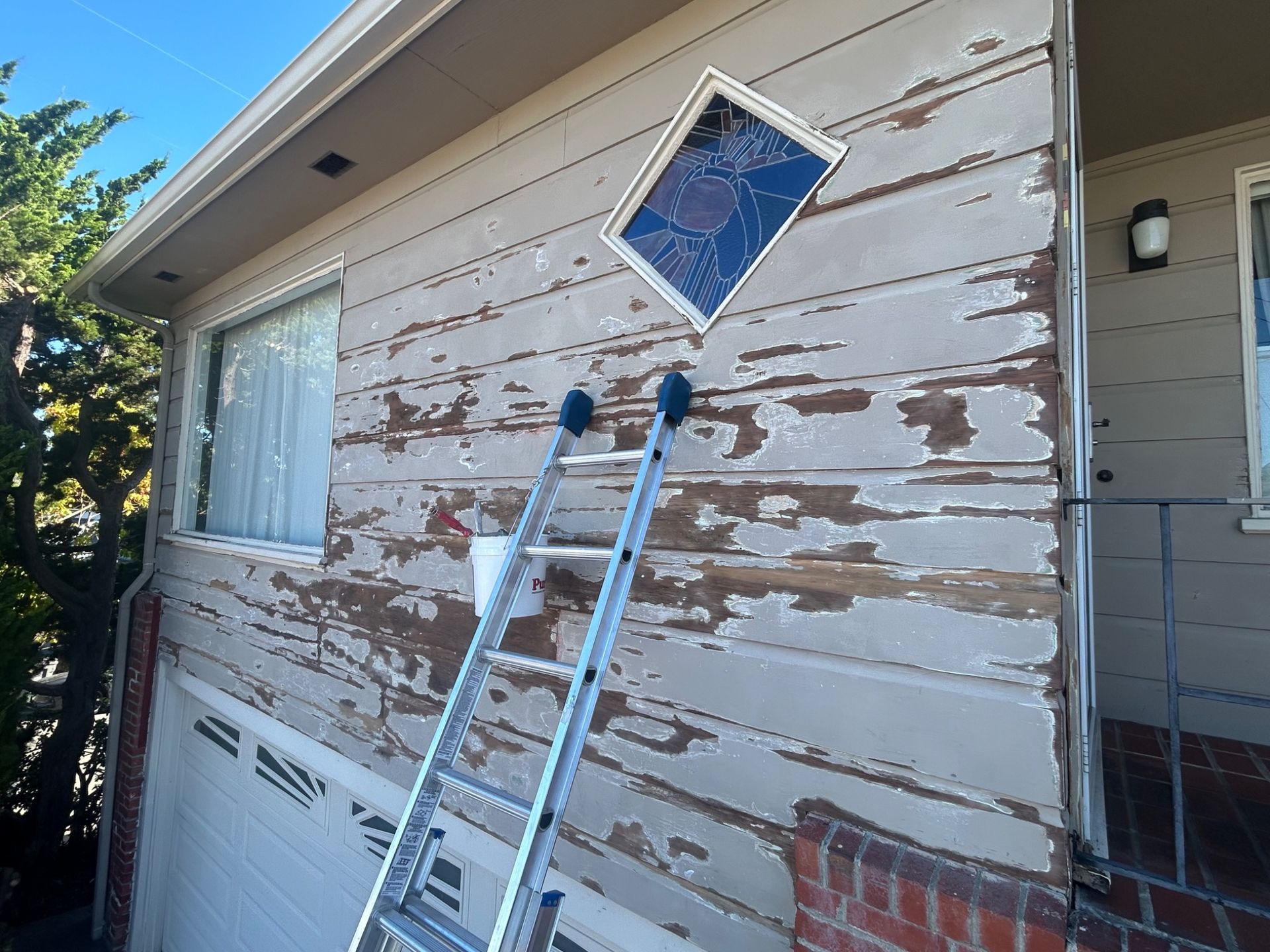 Ladder leaned against weathered house siding beneath a blue window, with peeling paint visible.