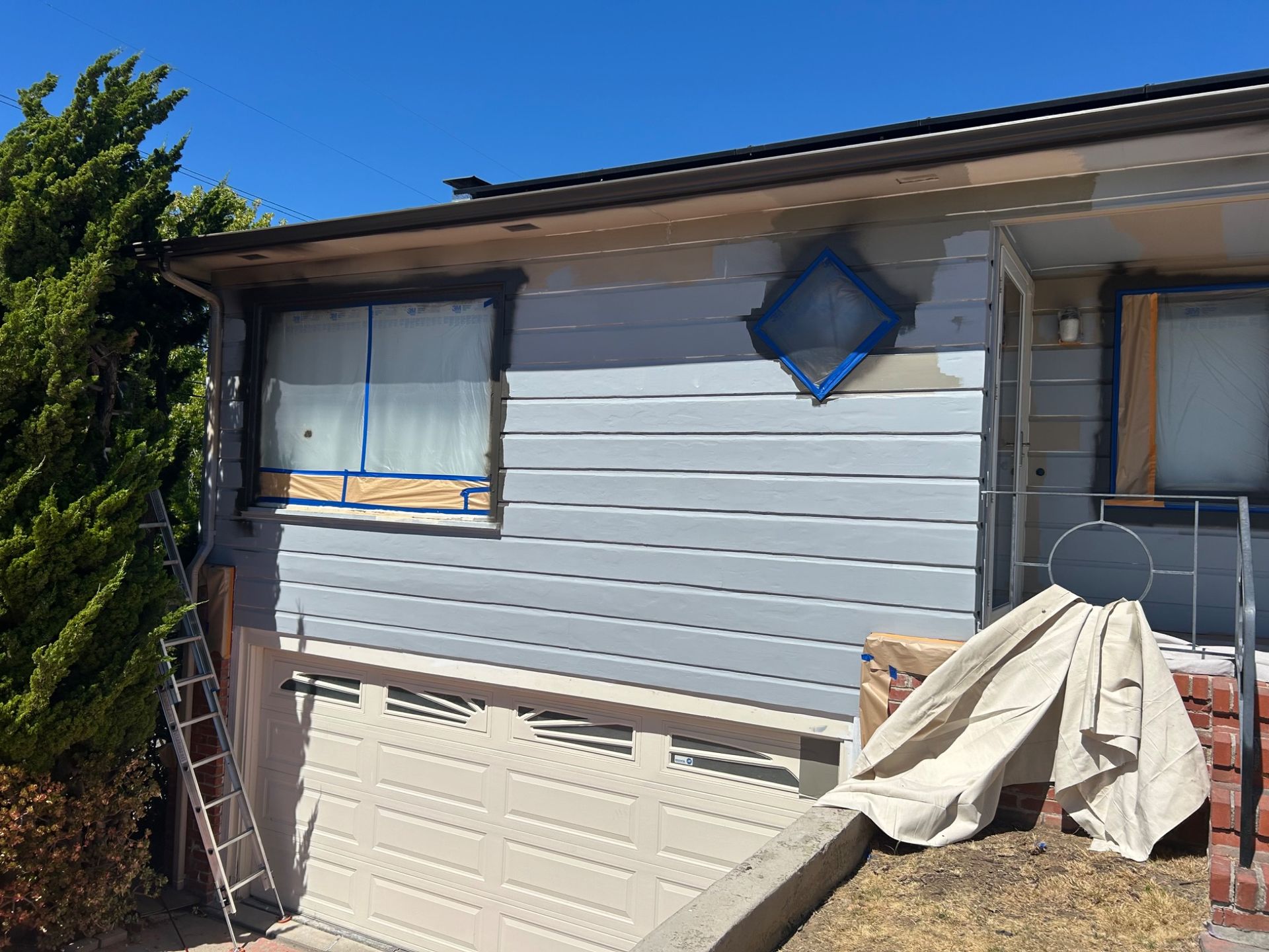 Small house exterior with blue-gray siding, a window, and covered items near the porch in bright sunlight