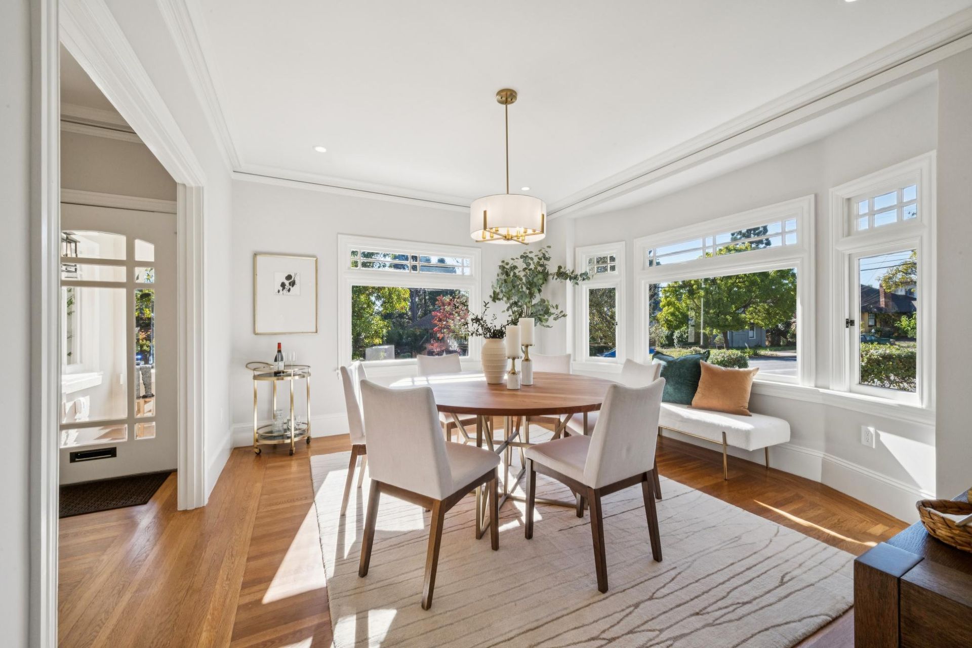 Bright dining room with round wooden table, white chairs, and large windows overlooking greenery