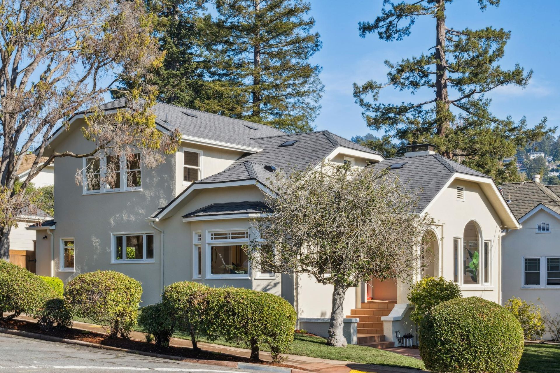 Two-story beige house with gray roof and front porch, surrounded by trees and shrubs