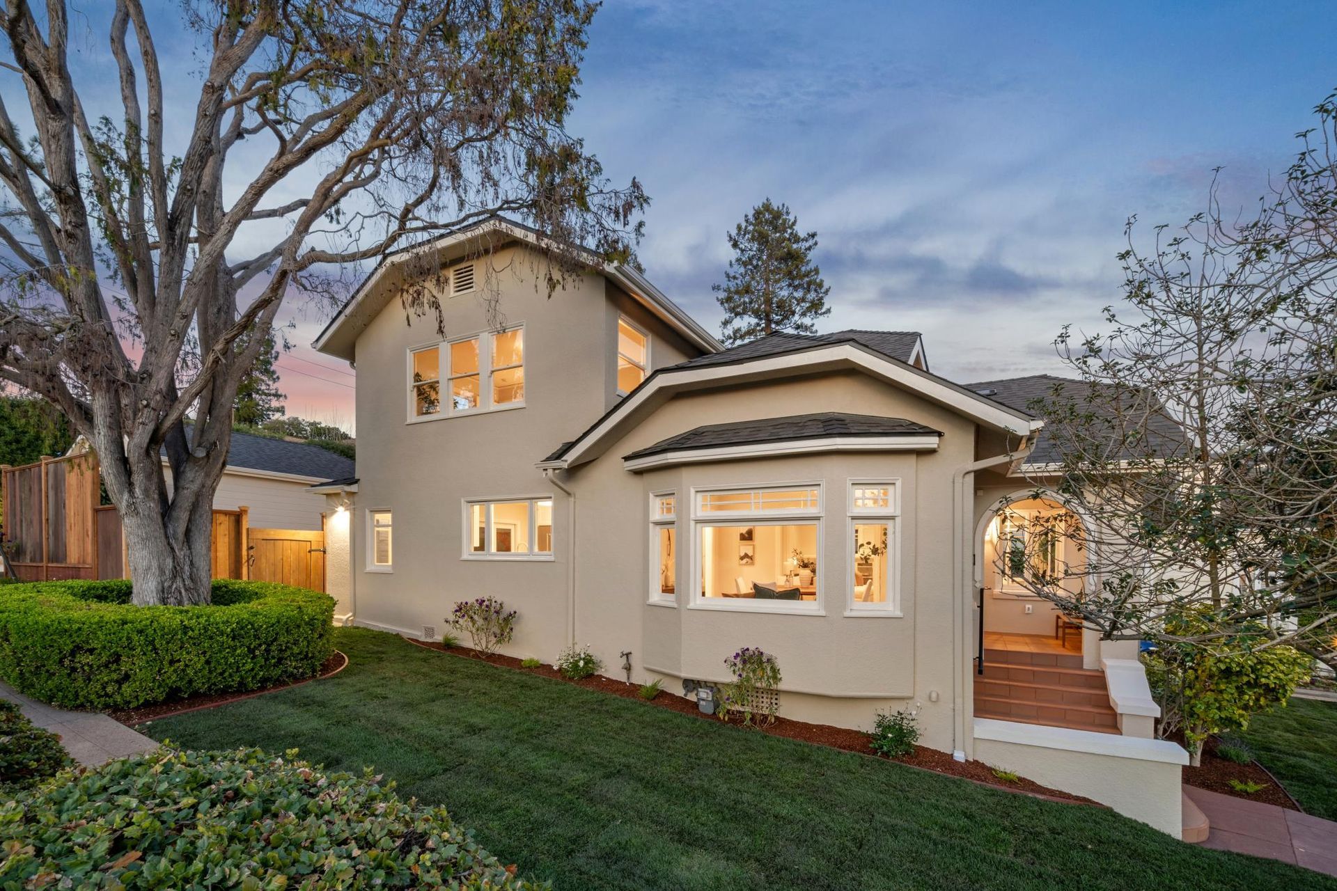 Two-story beige house with lit windows, a front yard, and trees at dusk