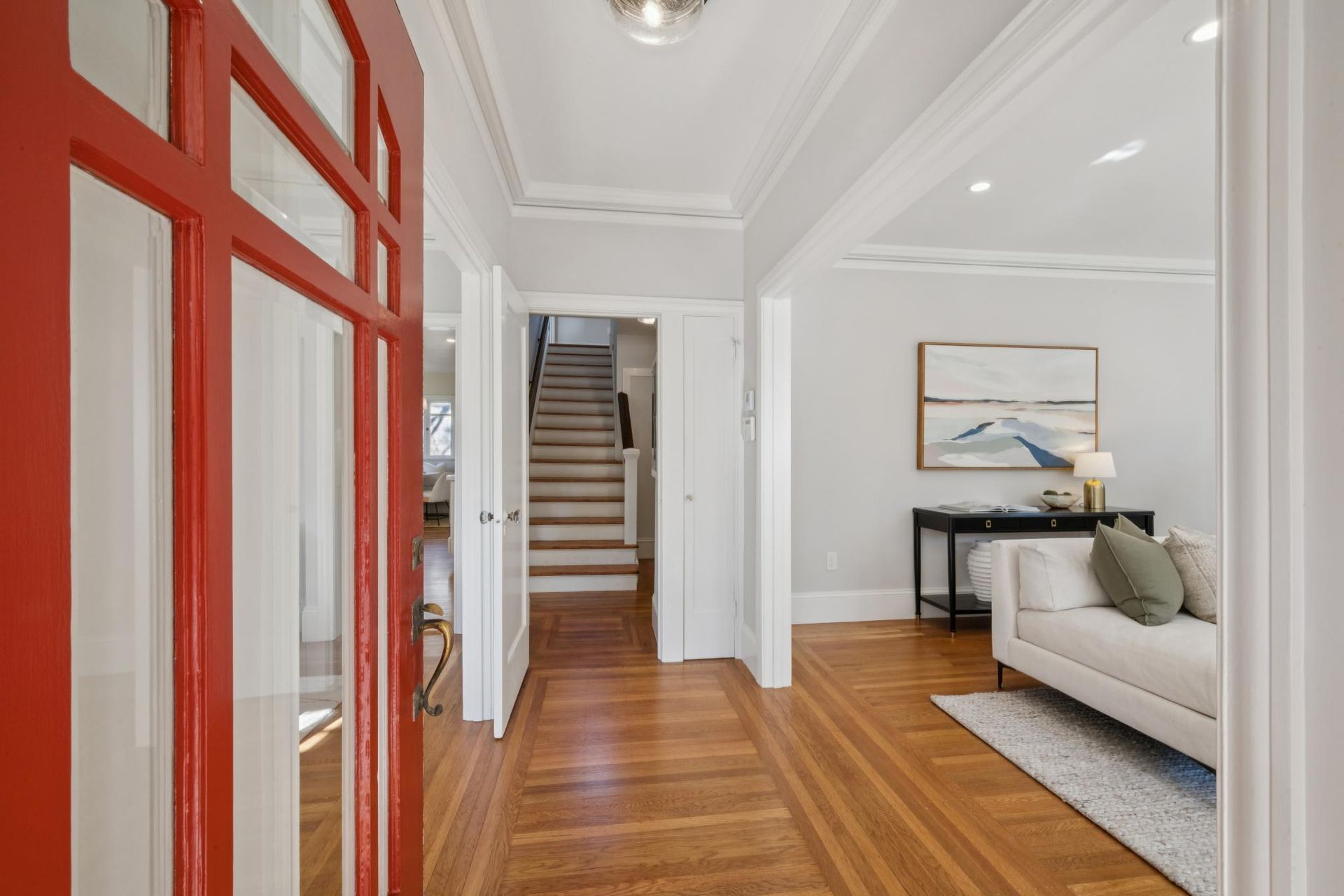 Bright hallway with a red front door, wood floors, staircase, and a glimpse of a living room.