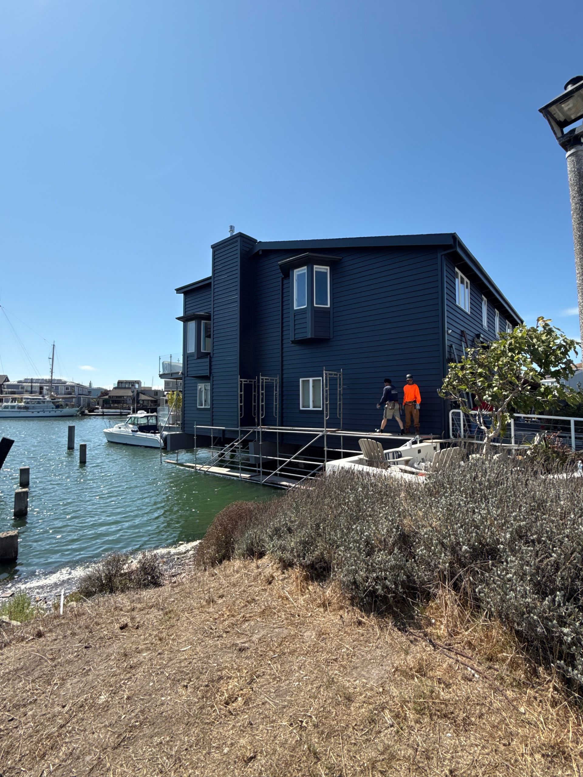 Dark blue houseboat on a canal beside a dock, with a person in an orange jacket under a clear sky.