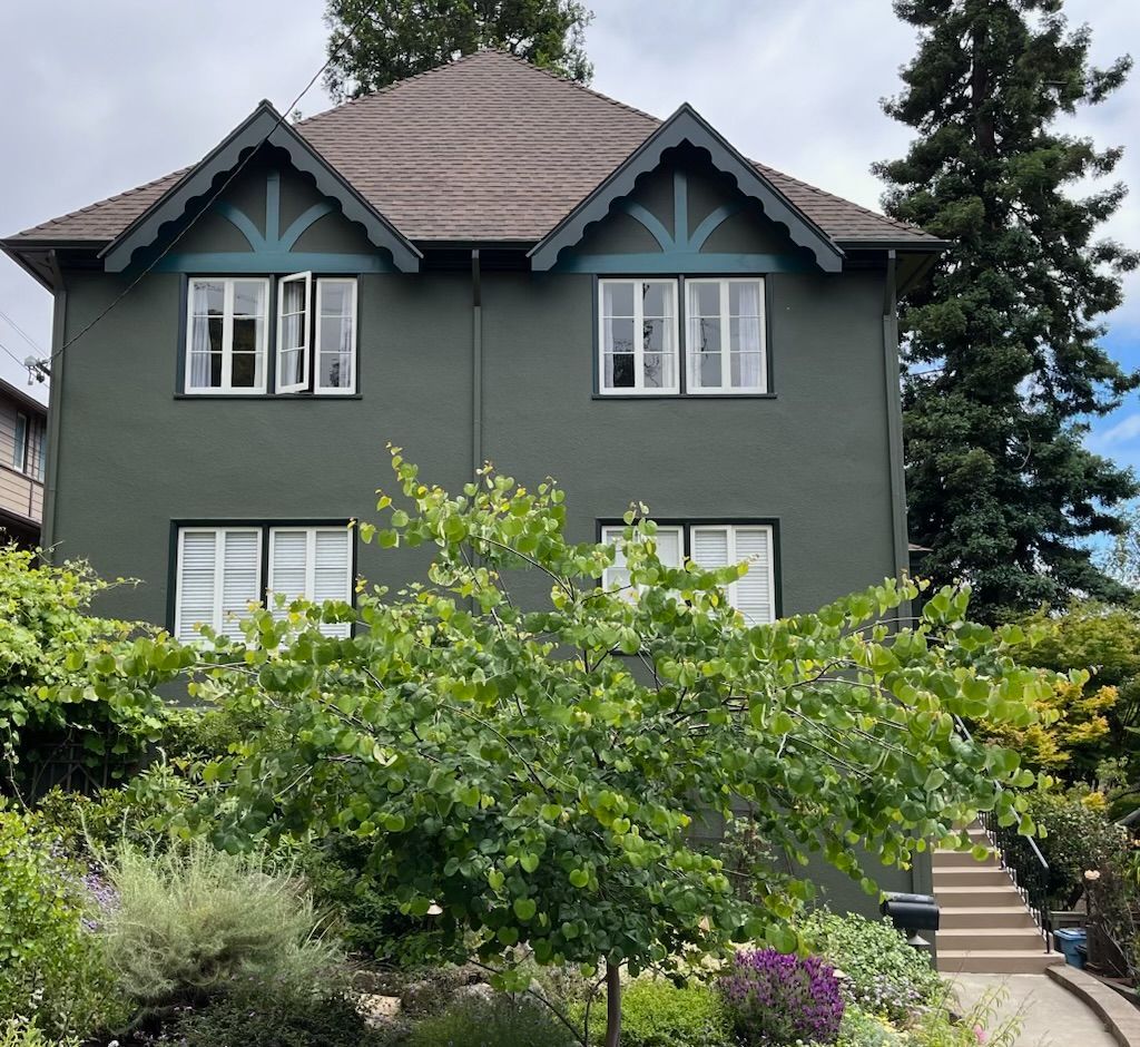Gray house with decorative gables, white-trimmed windows, and lush green shrubs in front