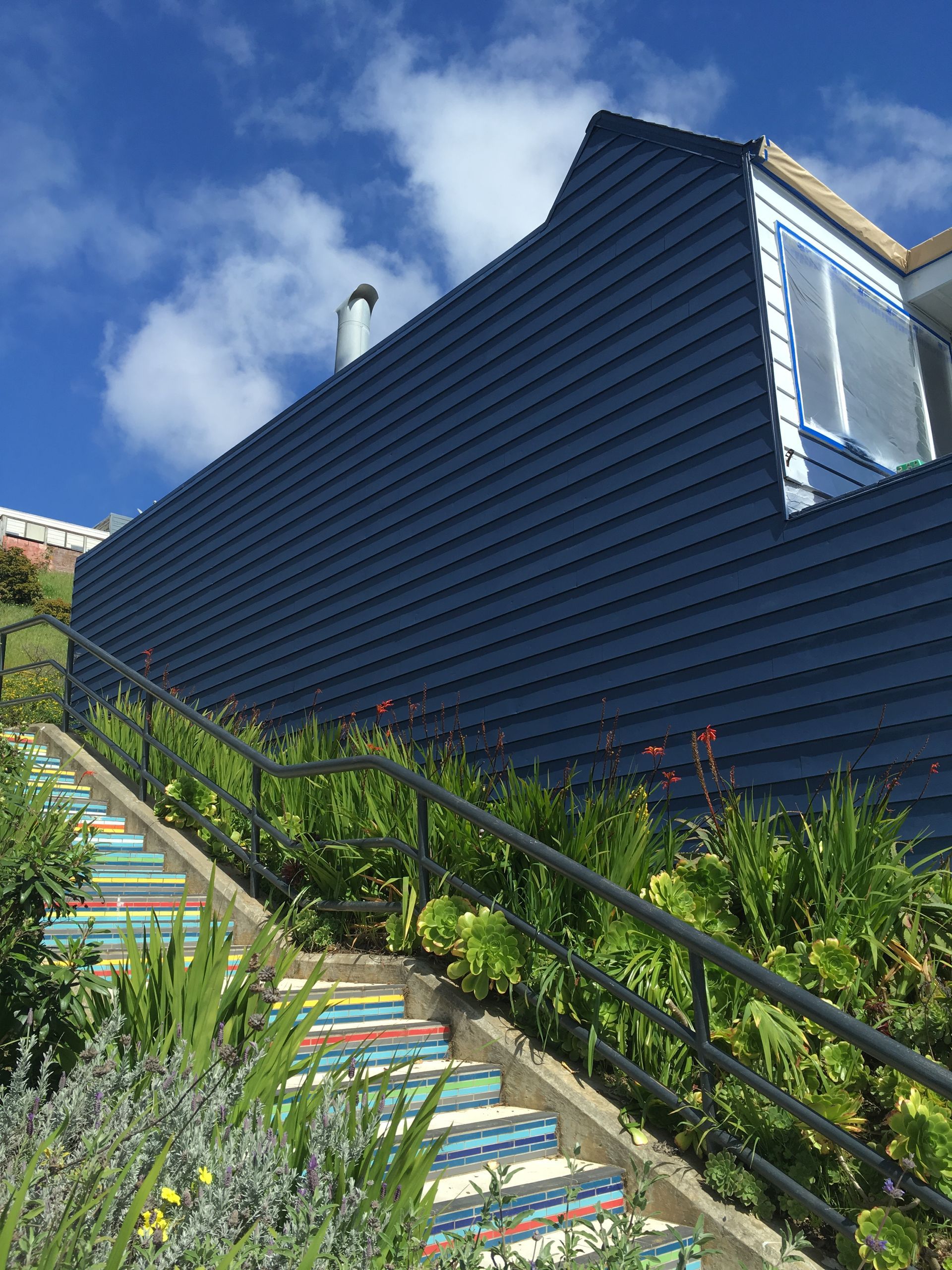 Blue-sided house with colorful stairs and lush greenery on a sunny hillside