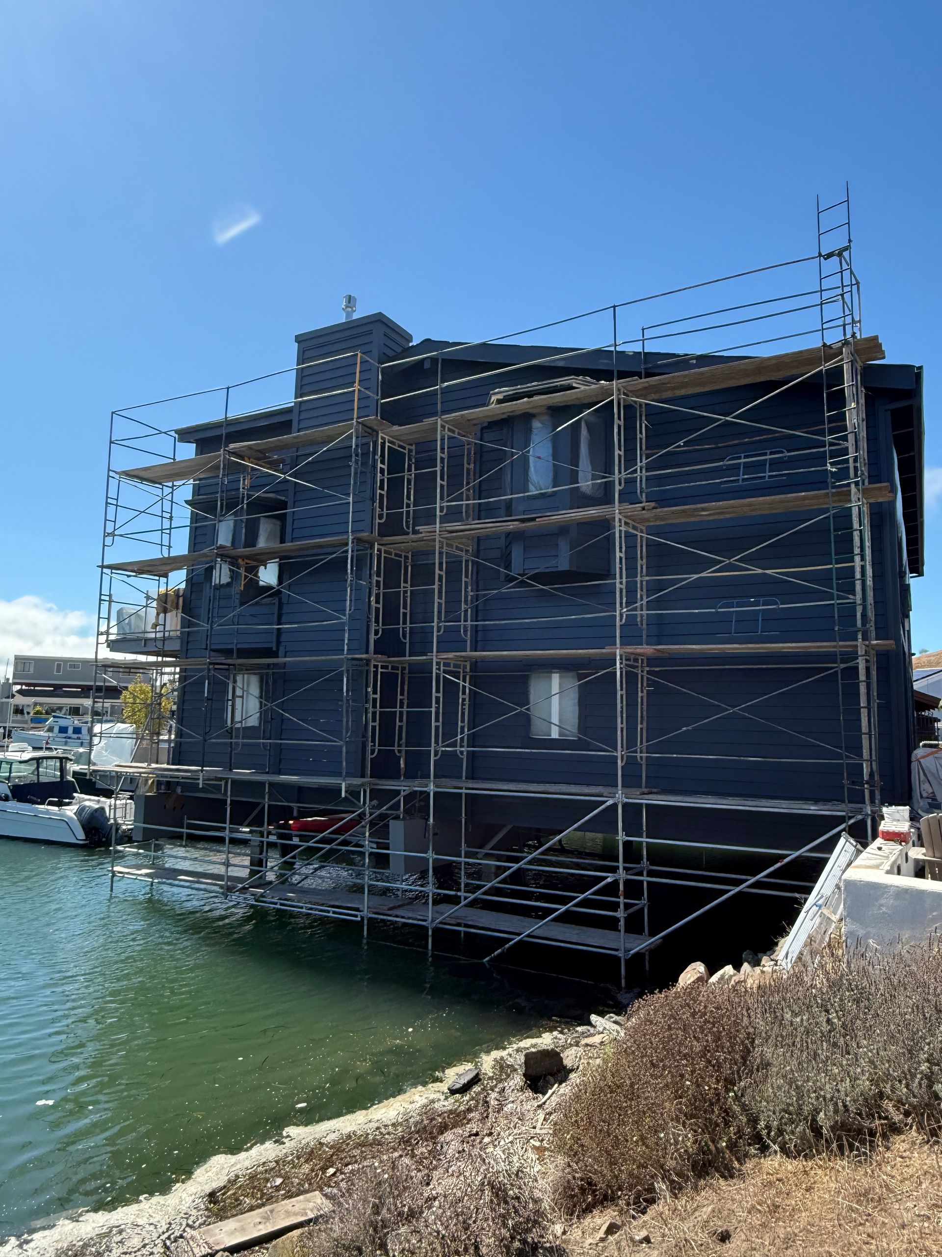Scaffolding covers a dark building beside calm water under a clear blue sky.