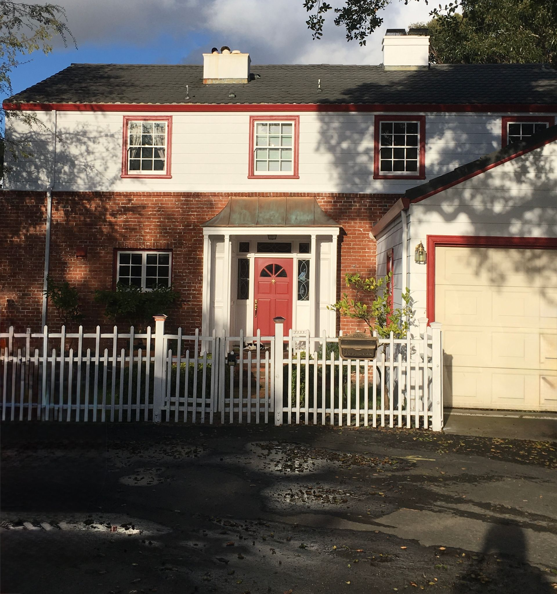 Two-story white house with red trim, a red front door, white fence, and attached garage in sunlight