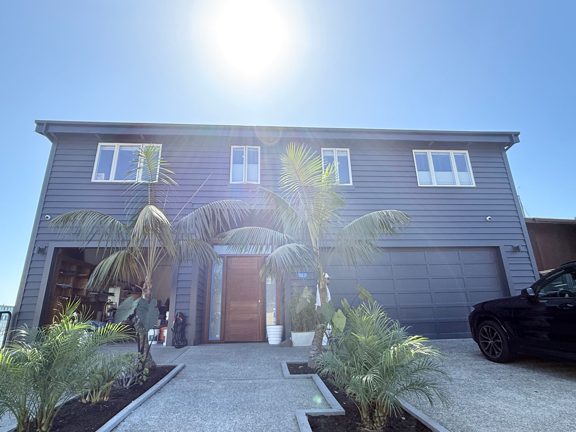 Two-story gray house with front walkway, palm trees, and garage under bright sunlight