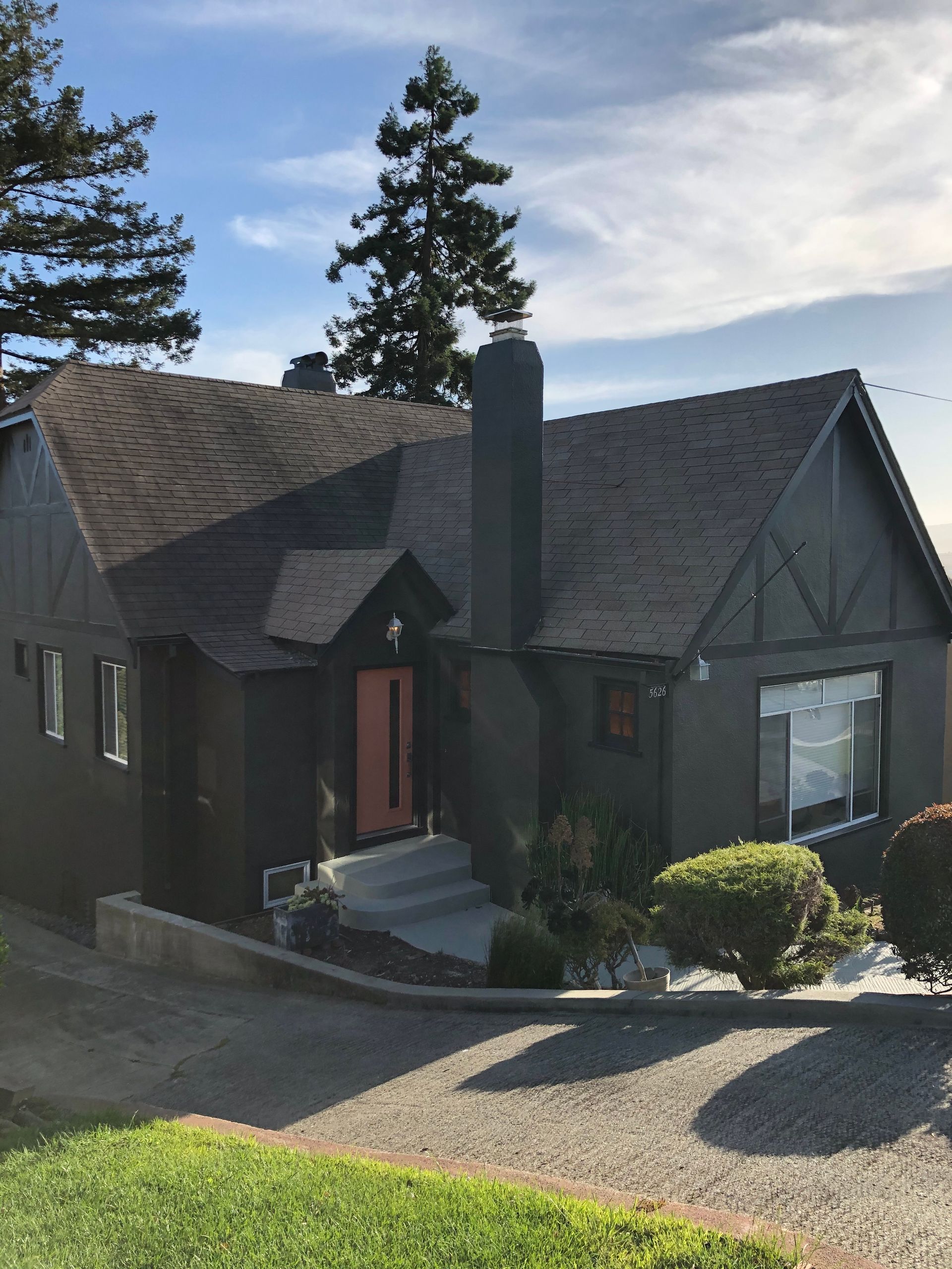 Dark gray house with a red front door, chimney, and gravel driveway, seen from the front under a blue sky.