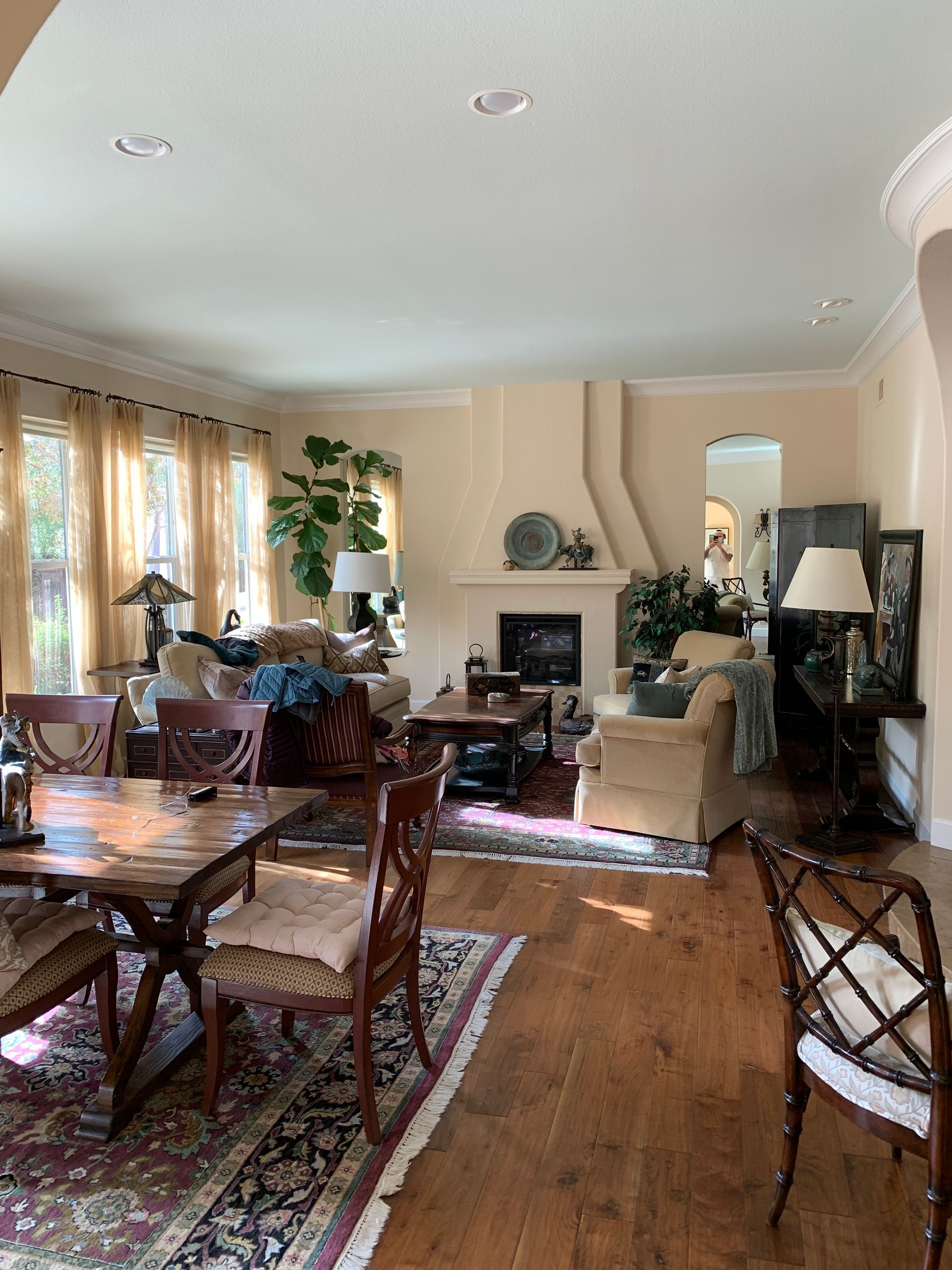 Bright living room with hardwood floors, patterned rugs, wooden chairs, a fireplace, and large windows with curtains