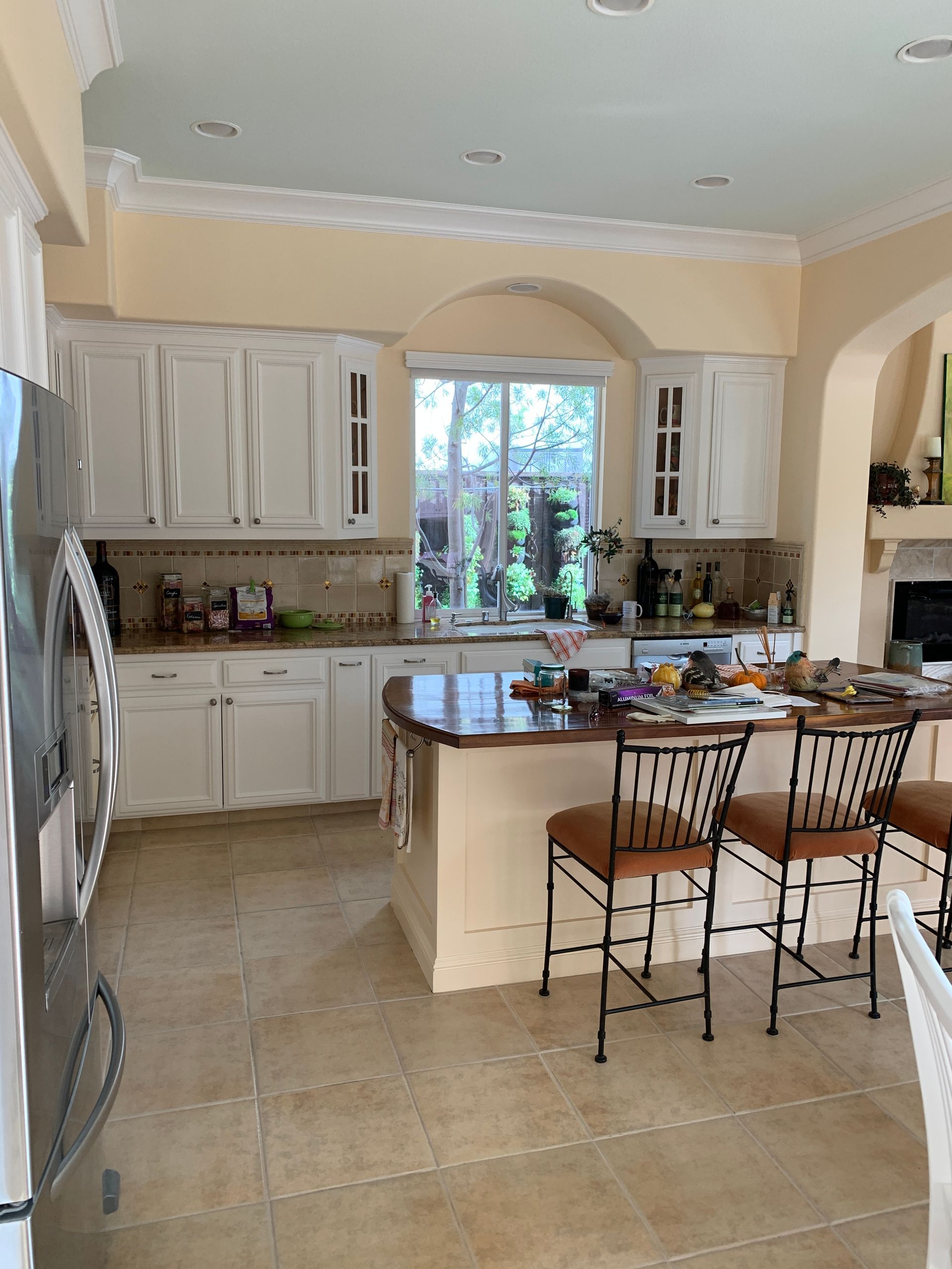 Bright kitchen with white cabinets, island, tiled floor, and two bar stools by a window.