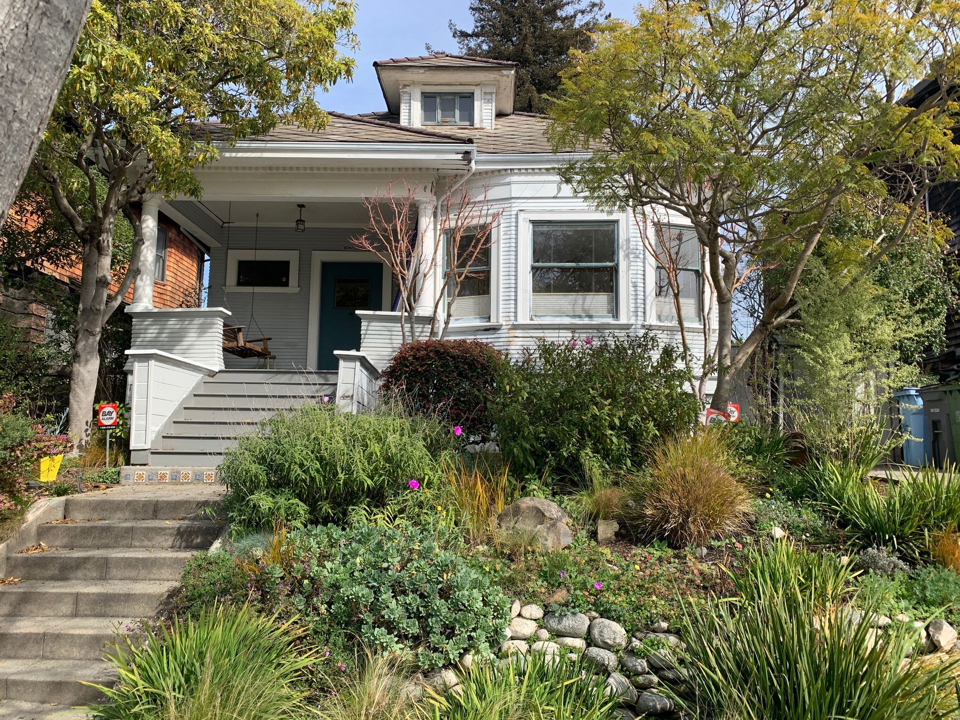 White house with front porch and steps, surrounded by lush garden and trees in autumn.