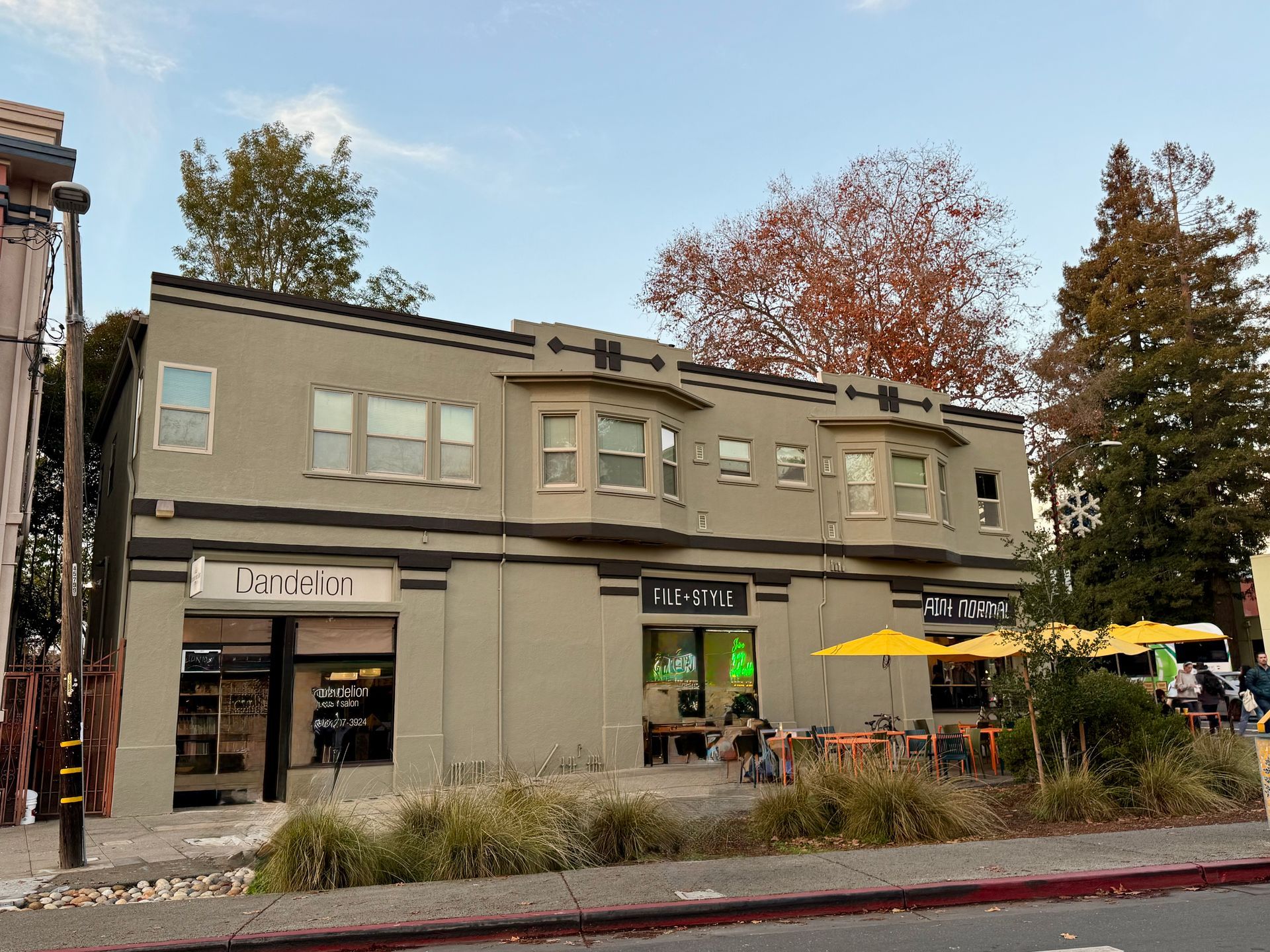 Street-level view of a beige storefront with outdoor seating, yellow umbrellas, and autumn trees behind it