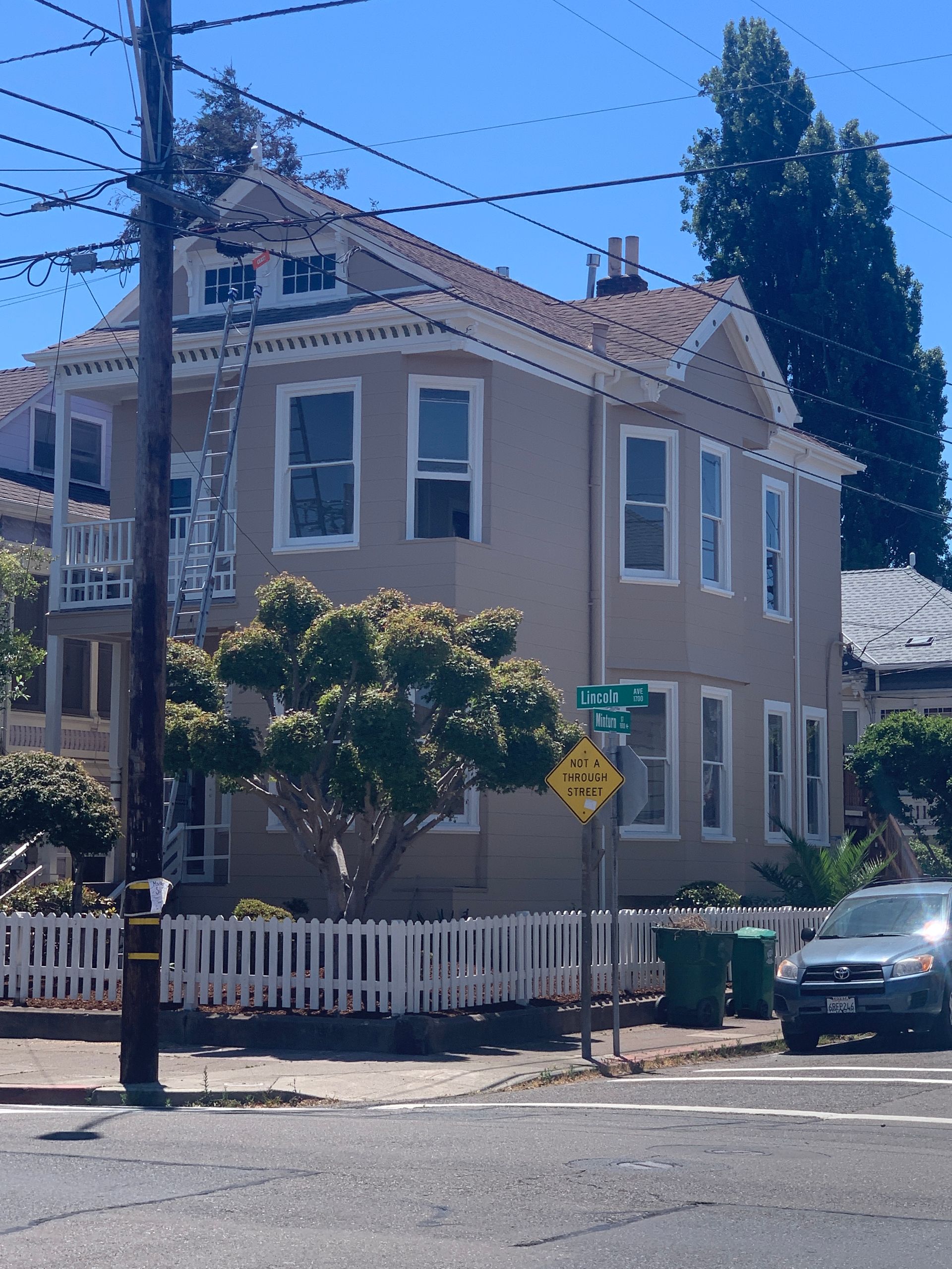 Two-story beige house on a street corner with white trim, a white fence, and a parked car beside it.