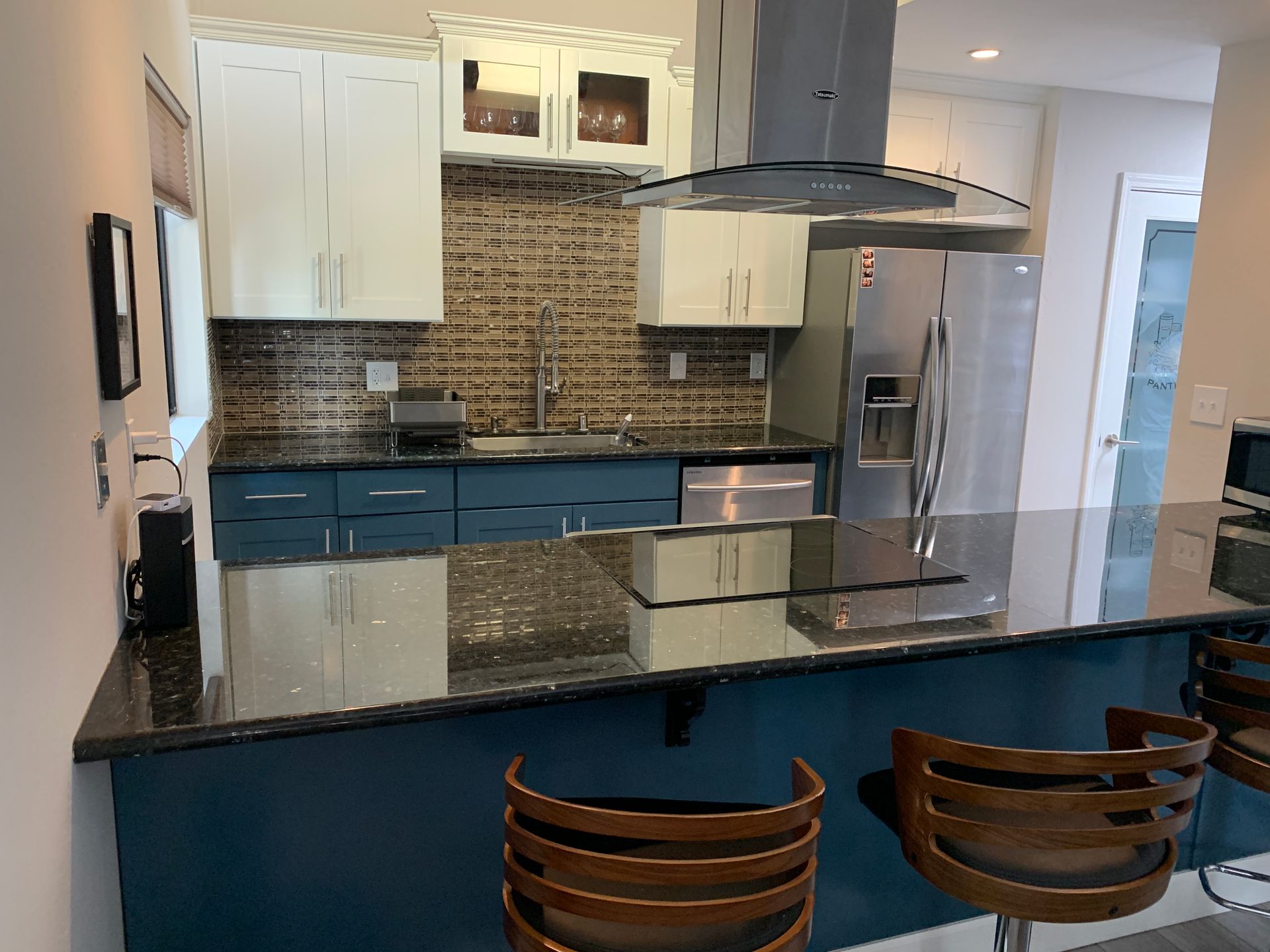 Modern kitchen with black granite island, blue lower cabinets, stainless range hood, and two wooden stools