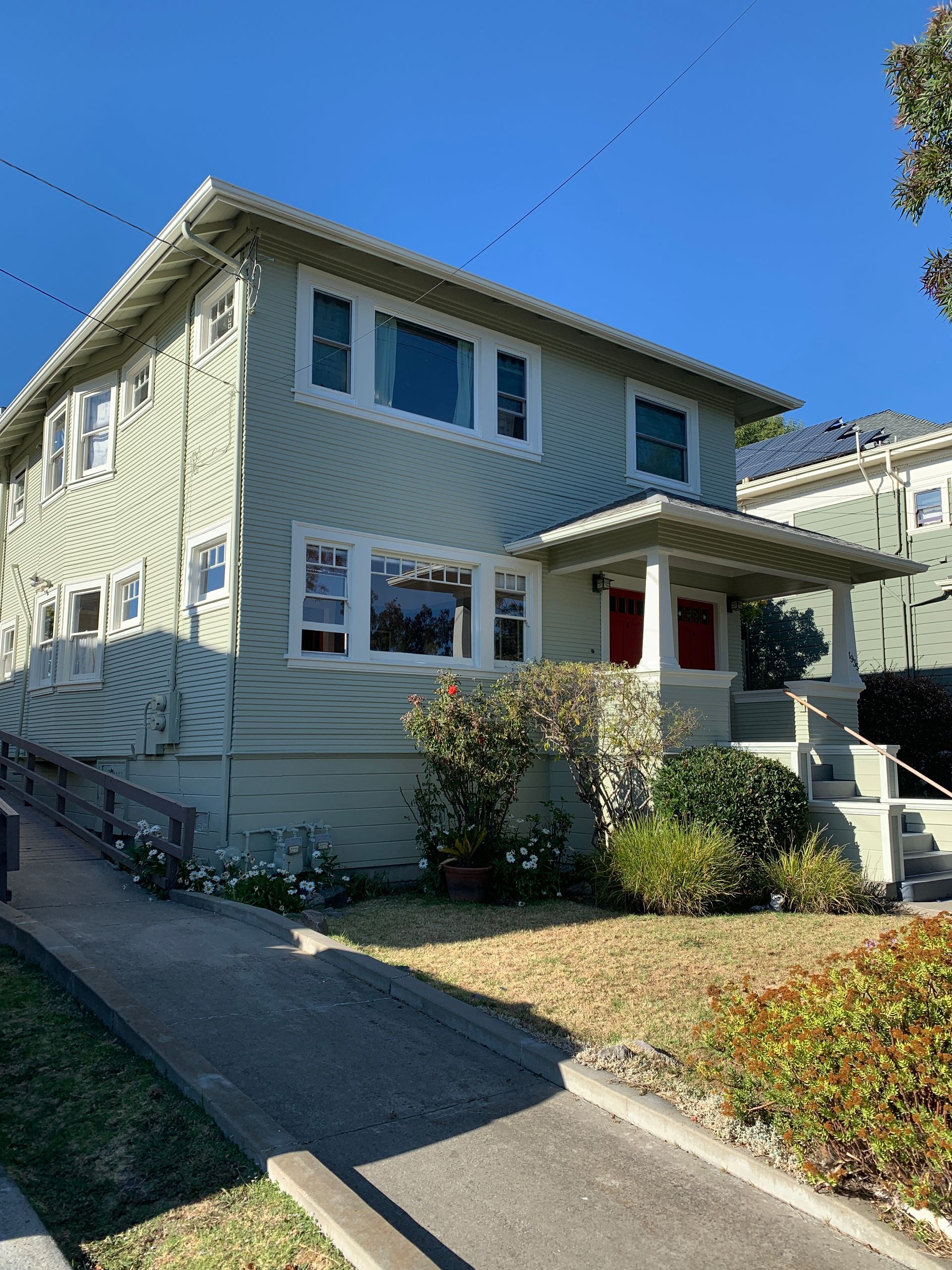 Two-story pale yellow house with a small front porch, white trim, and a sloped driveway under a clear blue sky.