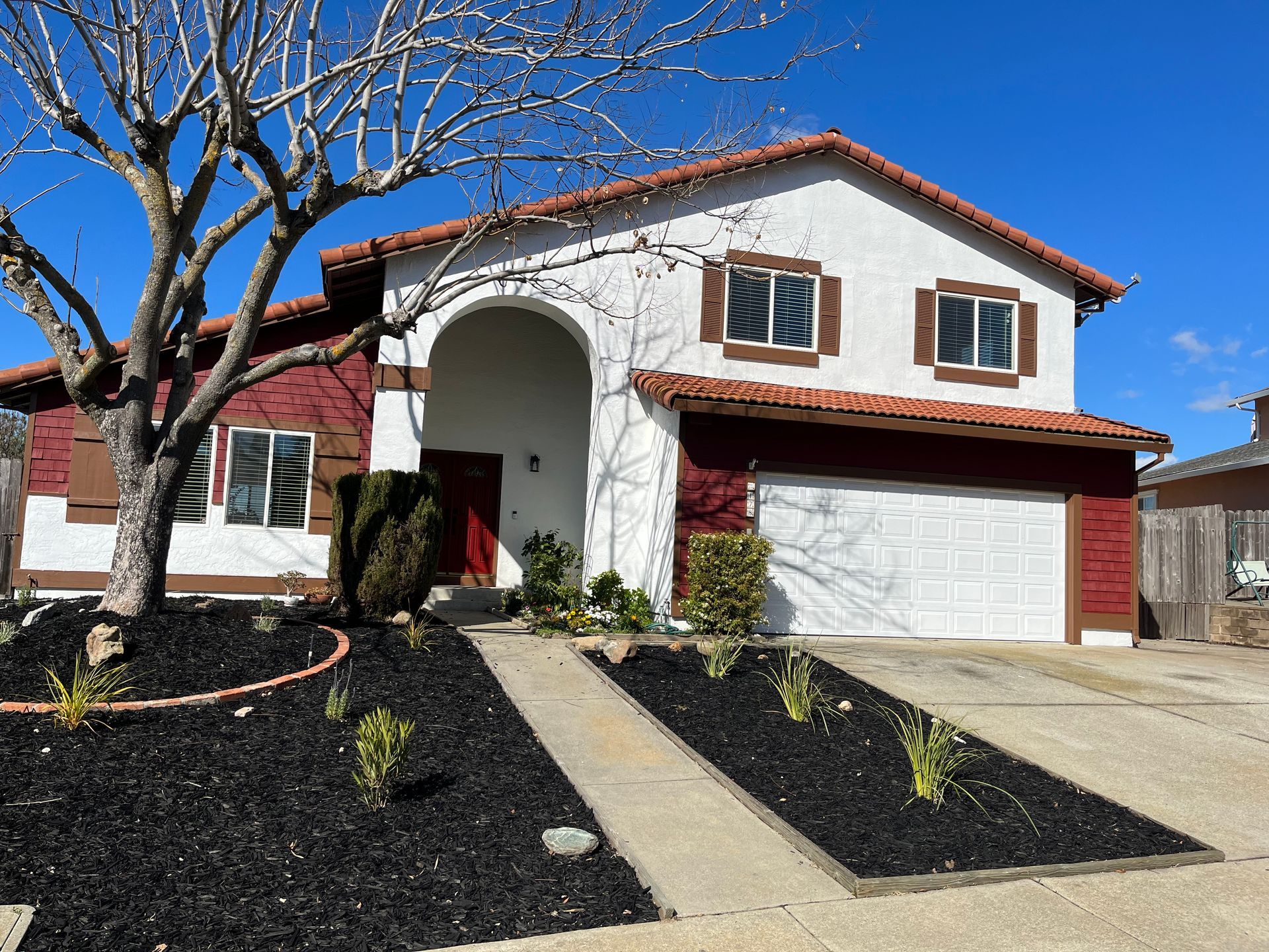 Suburban two-story house with red trim, white garage, front walkway, and leafless tree under a blue sky