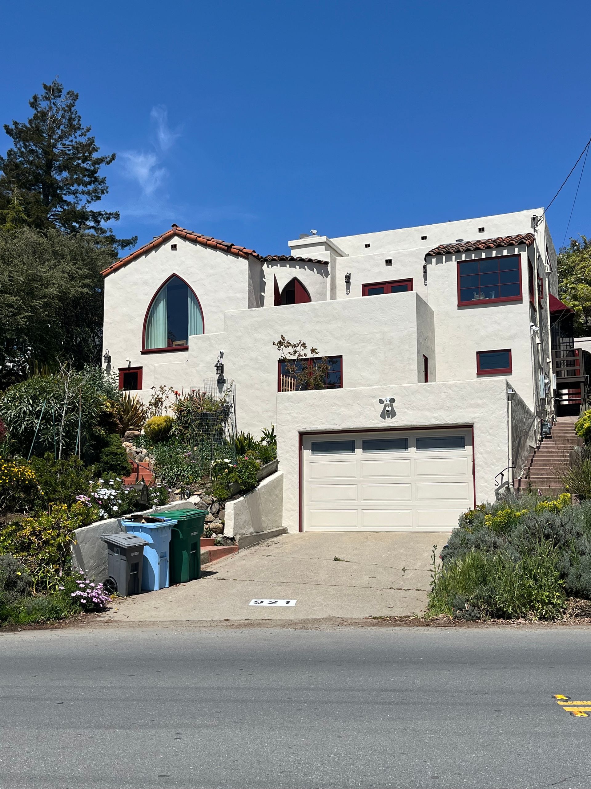 White stucco house with red tile roof, garage, and stairway on a hillside street under a clear blue sky