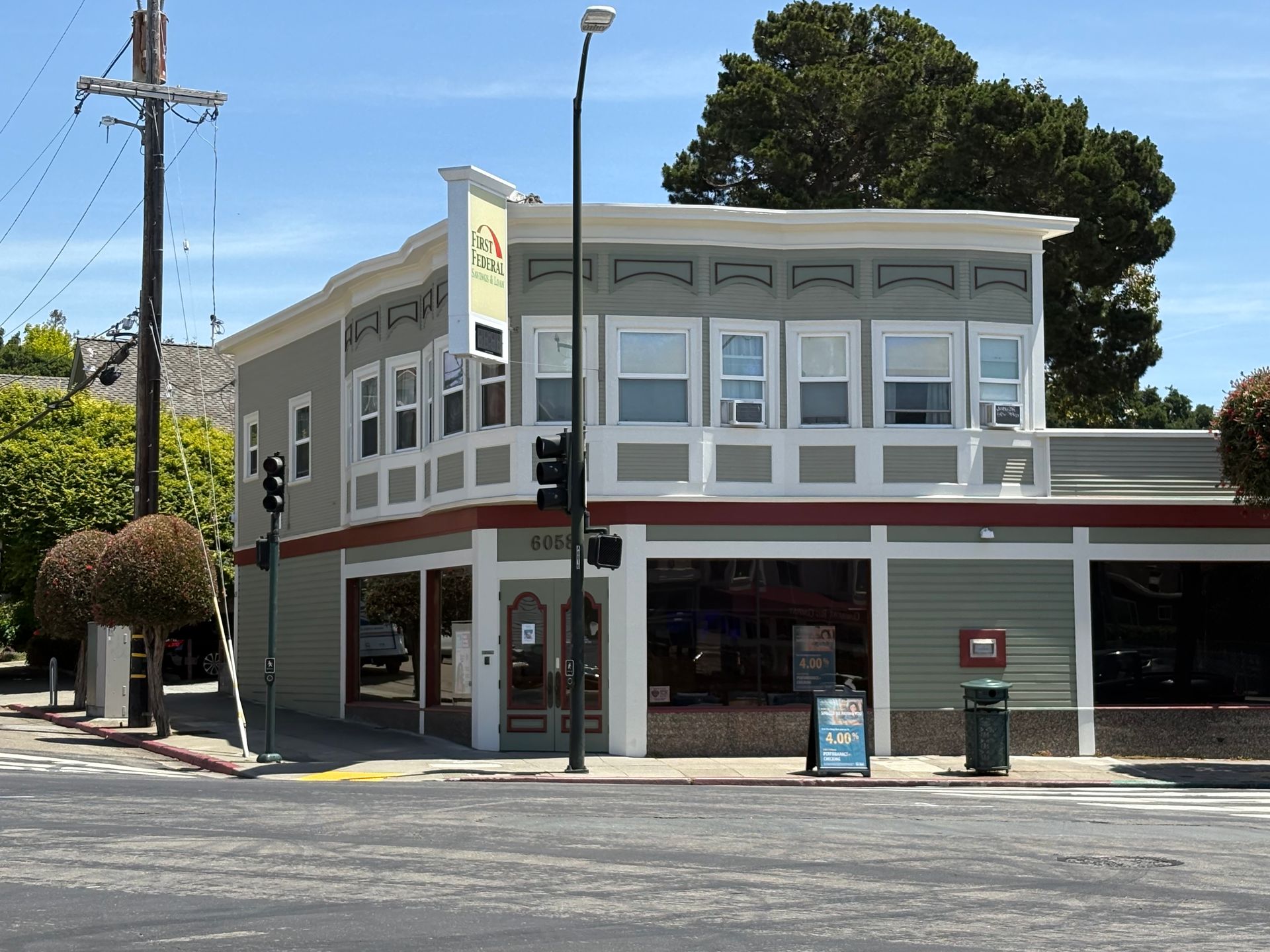 Corner storefront on a sunny street with white and gray facade, red trim, and a large tree nearby.