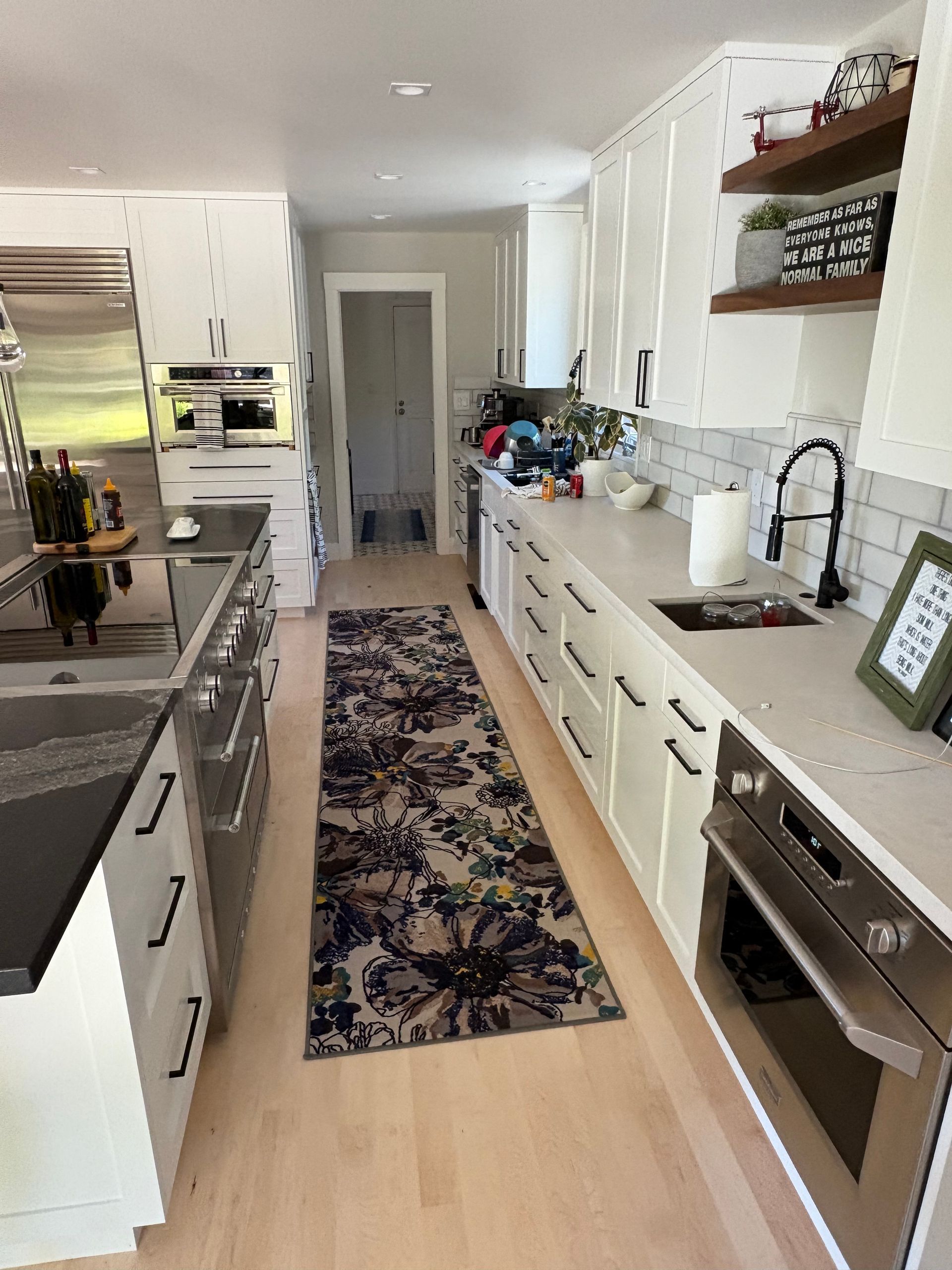 Bright galley kitchen with white cabinets, dark counters, a patterned runner, and stainless steel oven.