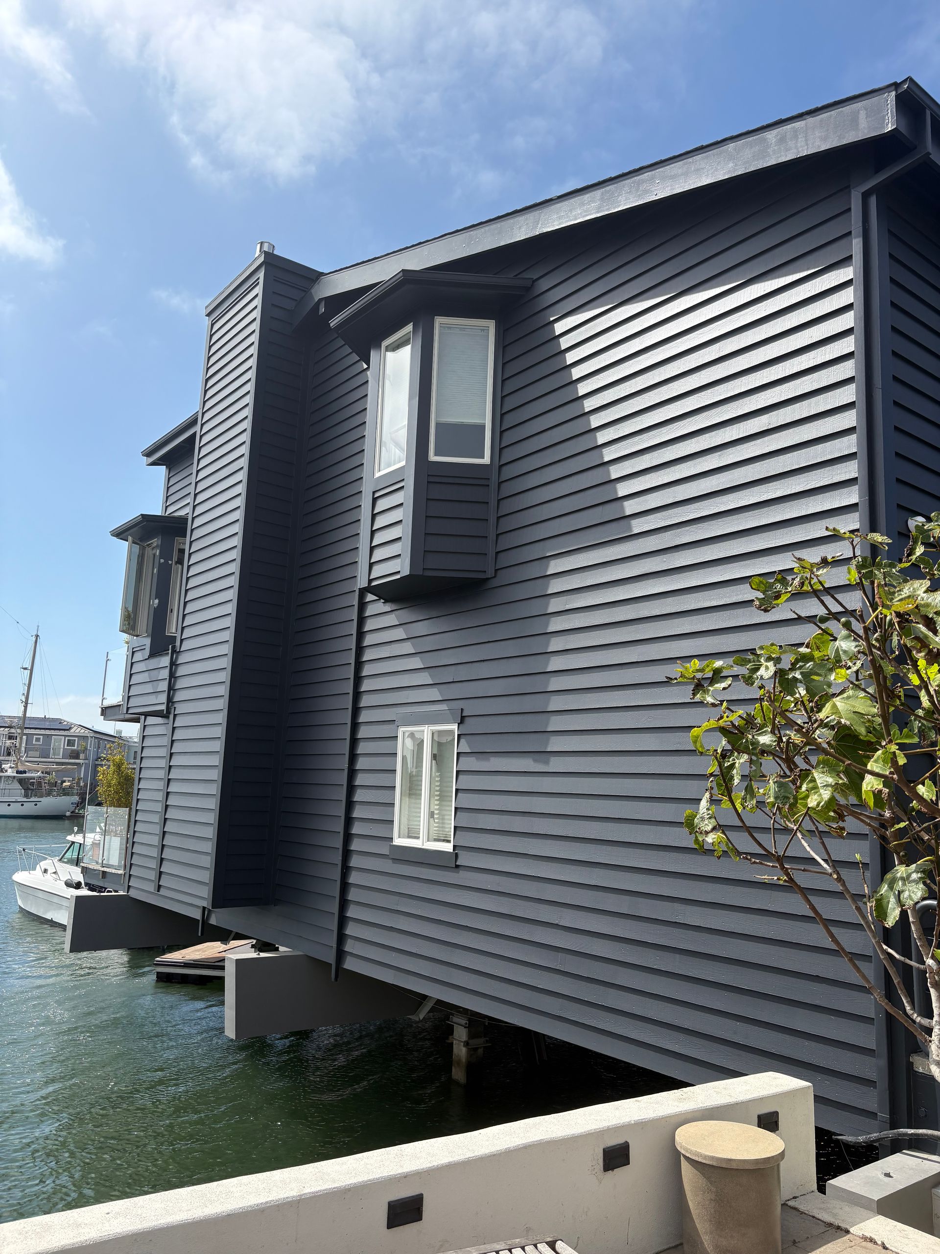 Dark gray waterfront building with bay windows and water below a bright sky