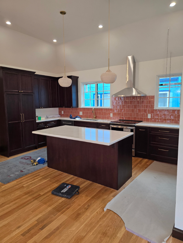 Modern kitchen with dark cabinets, white island countertop, brick backsplash, and hardwood floor being installed