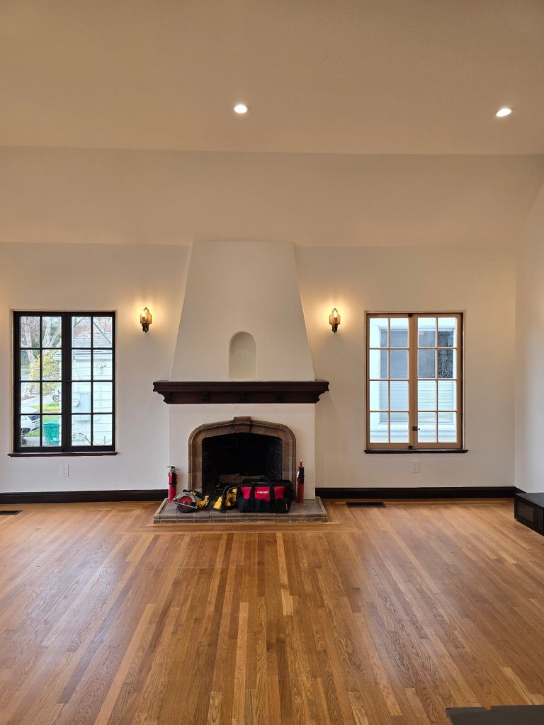 Empty living room with hardwood floors, white fireplace, and two windows on either side.