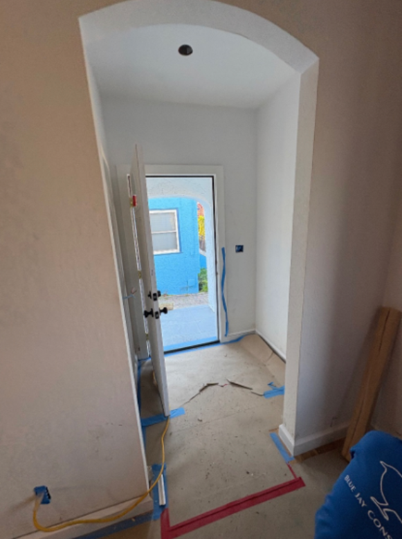 Narrow unfinished hallway with open door, blue wall outside, taped floor, and tools along the walls.