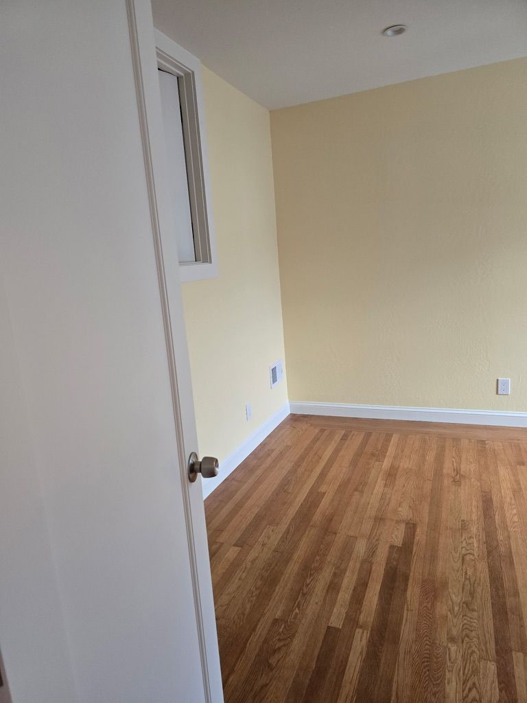 Empty beige room with wood floor, open door, and small high window.