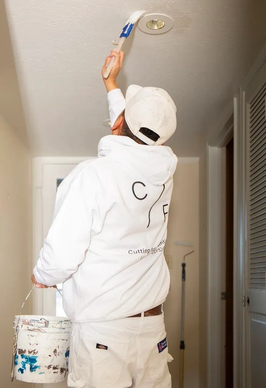 Cutting Edge Painting painter in white work clothes painting a ceiling with a roller beside a paint bucket.