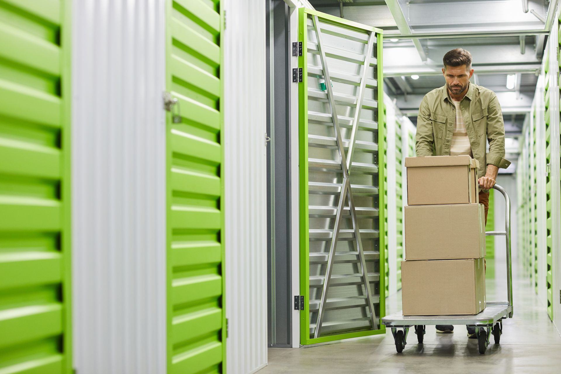 Man rolls a cart of stacked cardboard boxes past rows of green and white indoor storage unit doors.