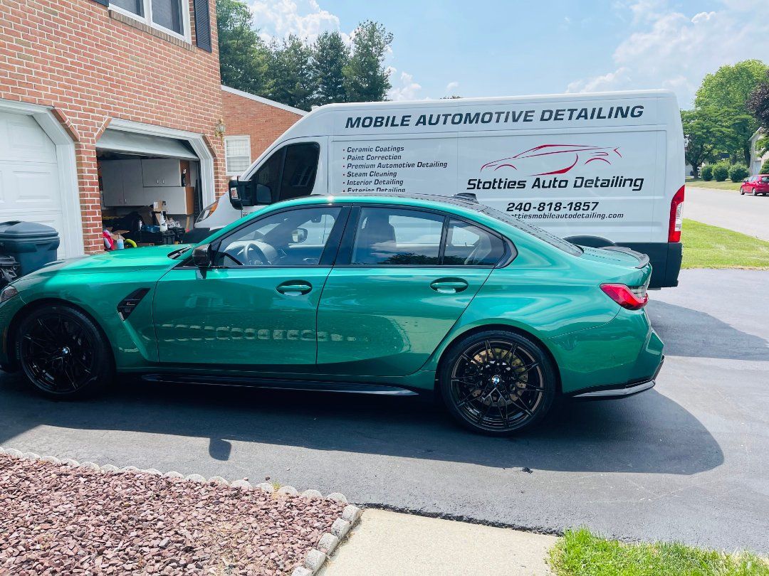 Green sedan parked in a driveway beside a white auto-detailing van and brick house