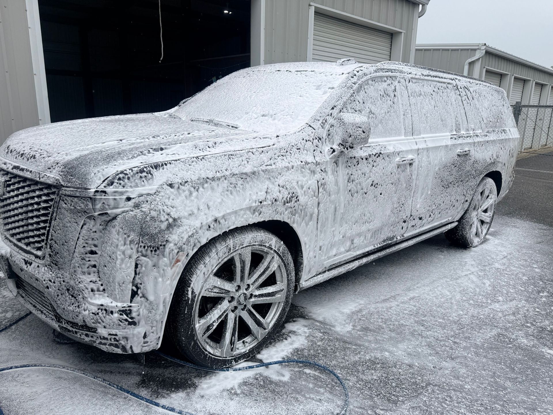 A large SUV covered in white foam, undergoing a car wash. It's parked outside a garage.