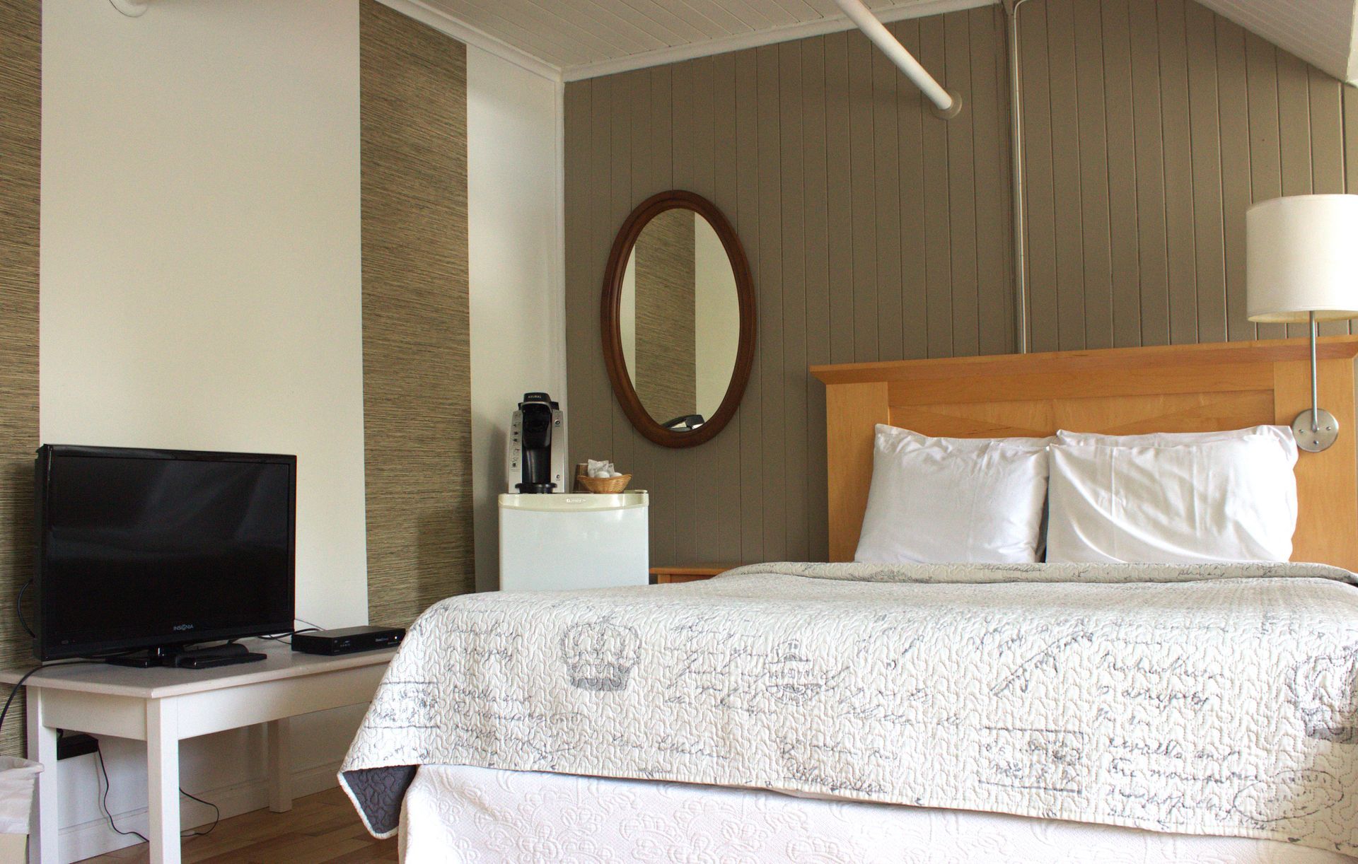 Bedroom with white bedspread, wooden dresser, chair, and lamp in a simple, bright room